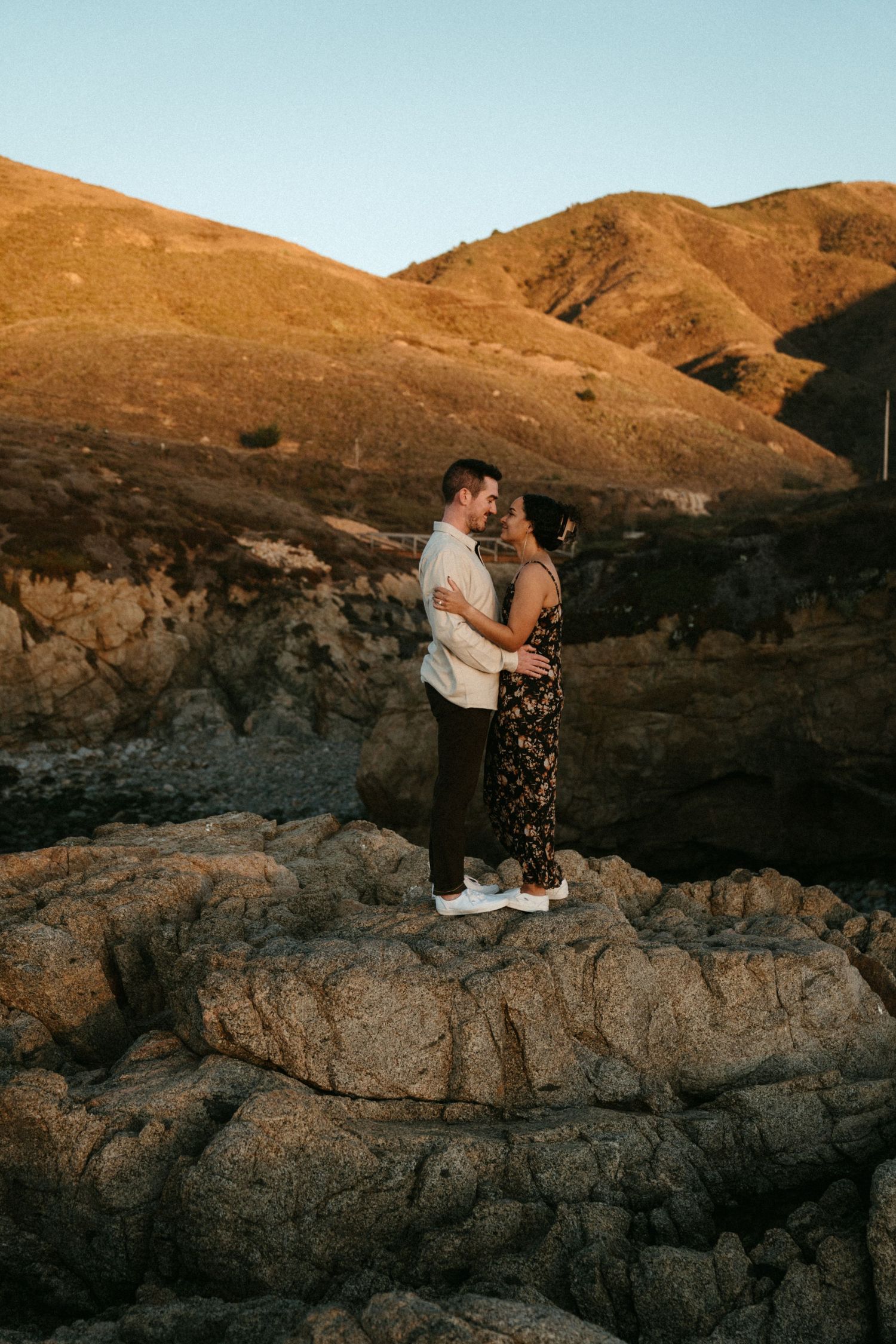 A couple embraces on rugged rocky terrain against golden rolling hills during sunset in a desert landscape.