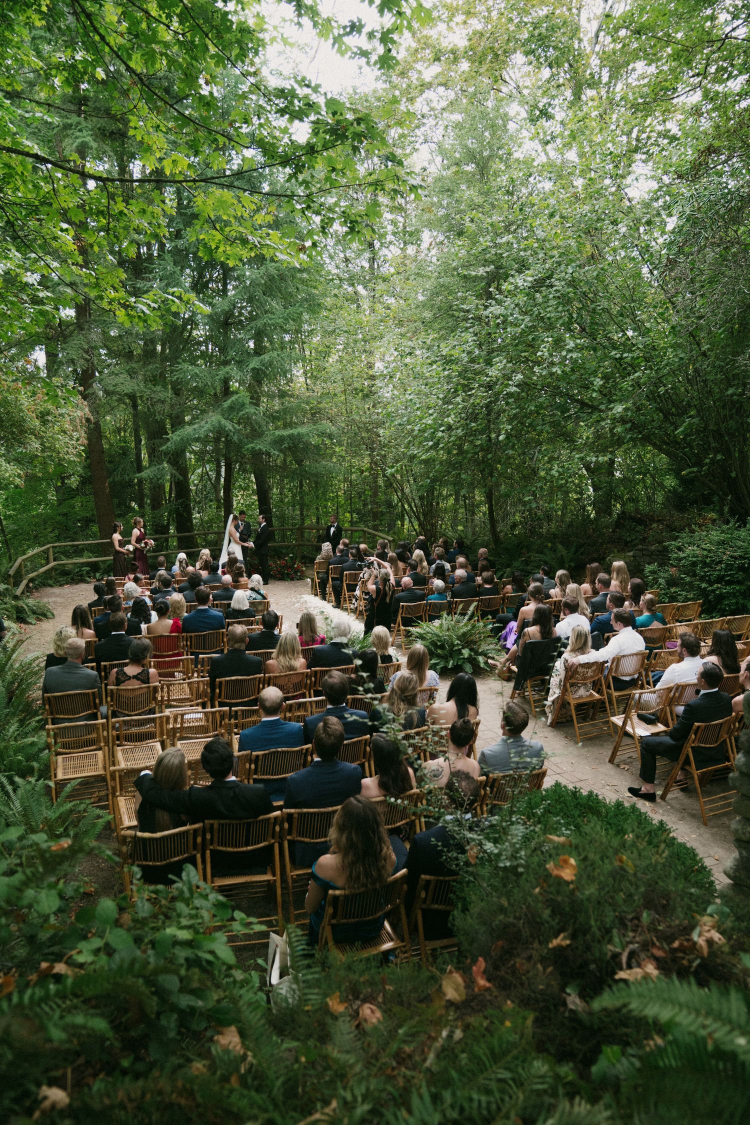 An outdoor wedding ceremony in a lush forest setting with wooden benches filled with guests.