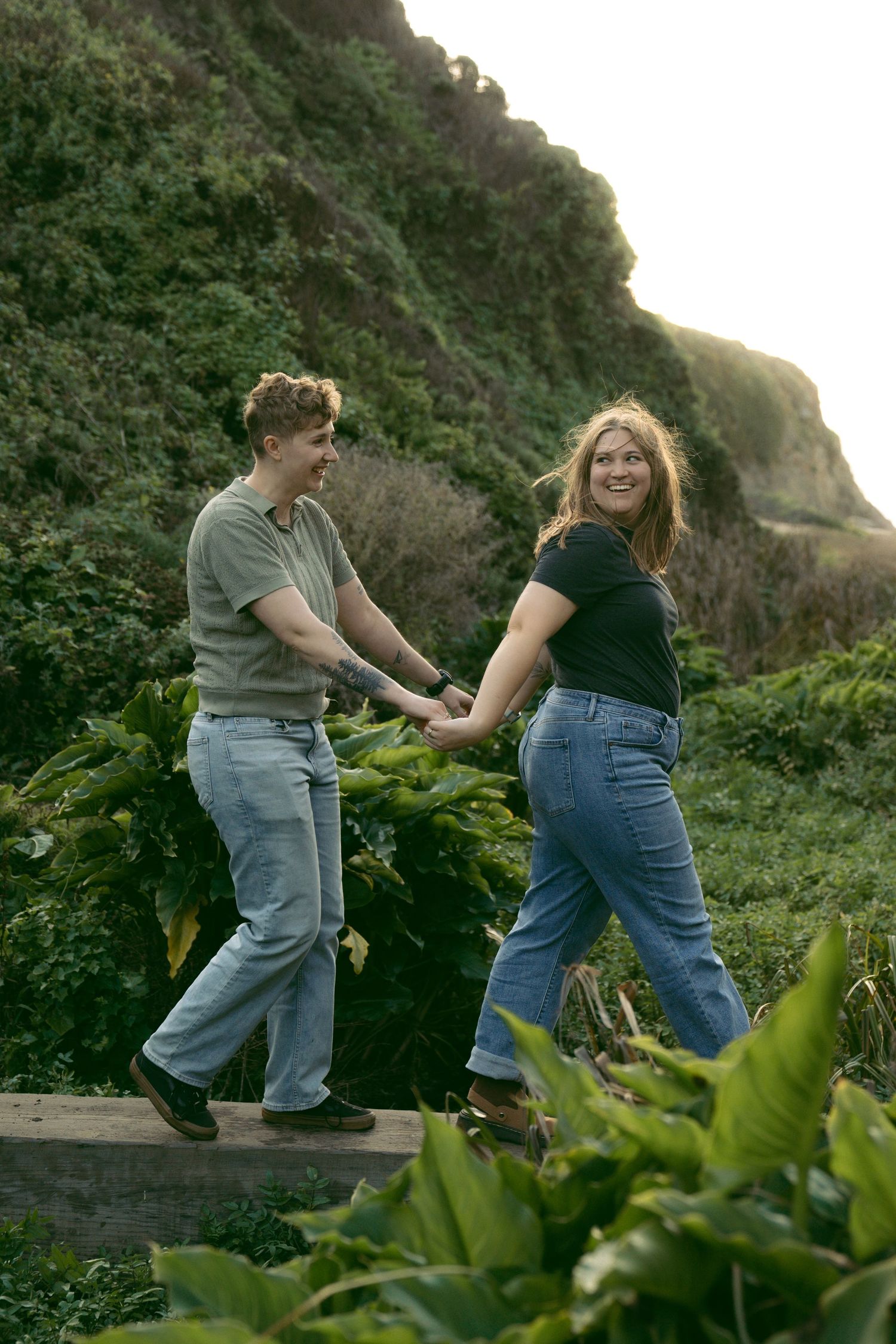 Two people playfully dance together on a scenic hillside path with green plants in the foreground.
