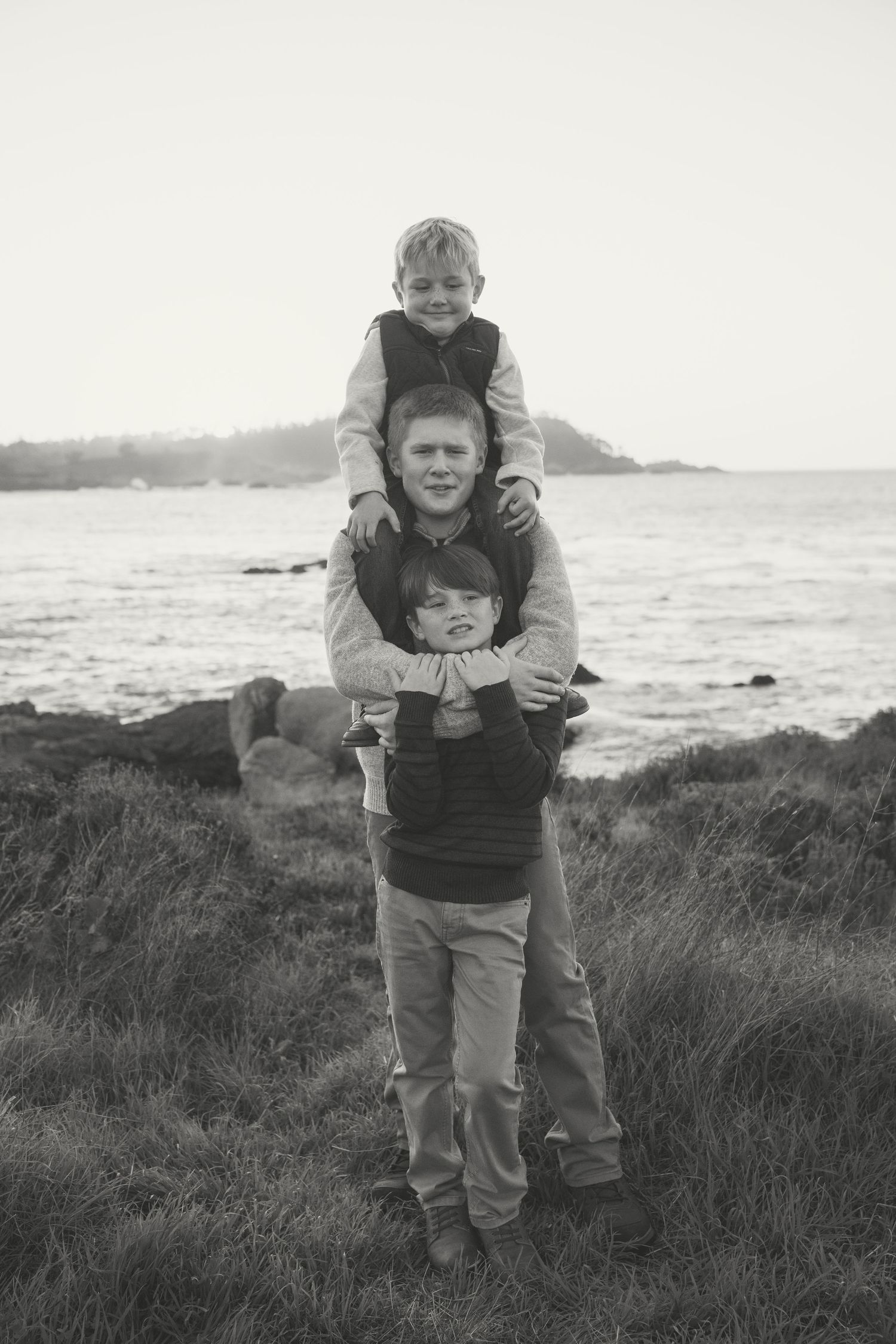 Black and white photo series showing two people enjoying a playful piggyback ride on a grassy coastal hillside.