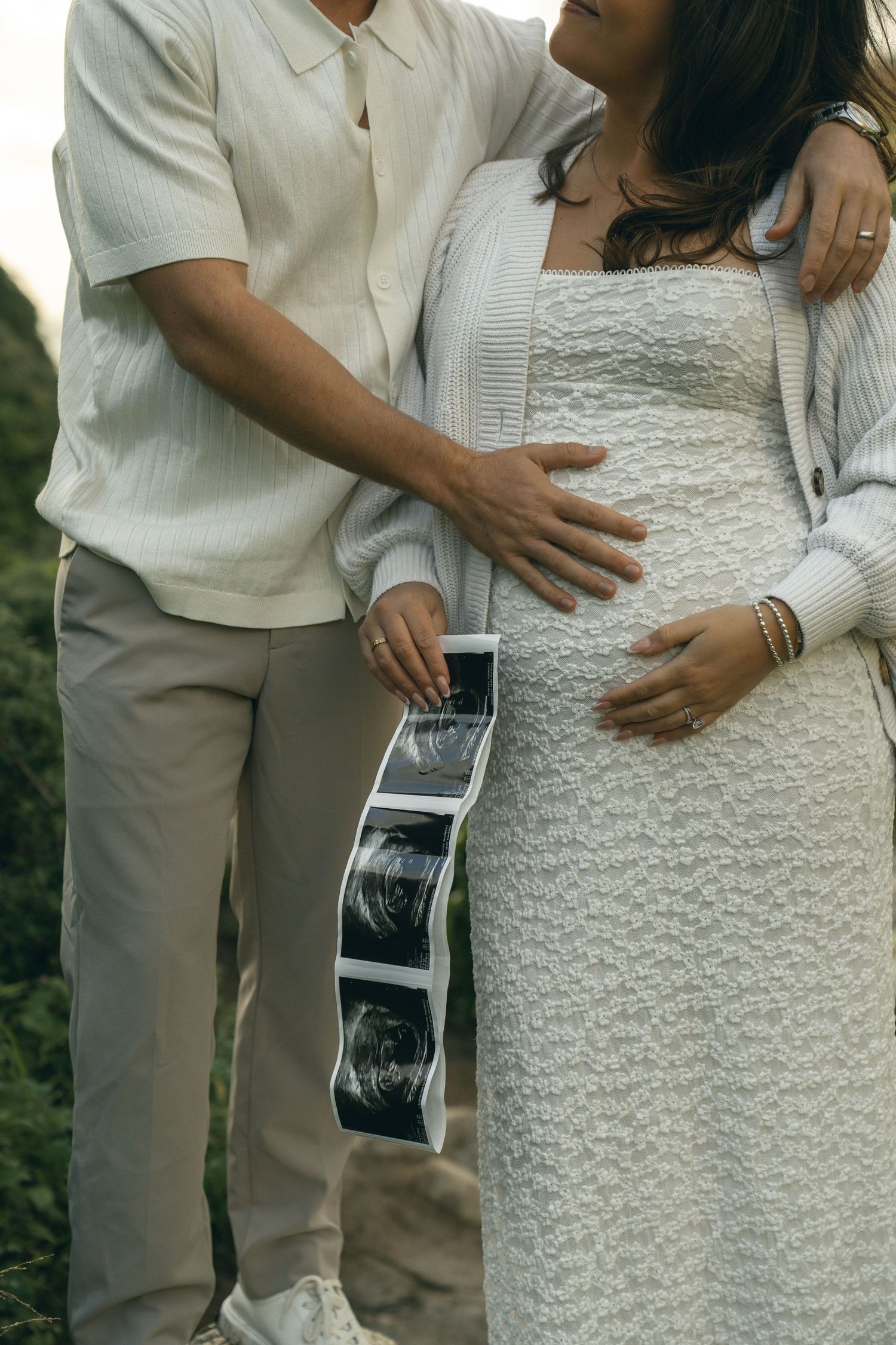 Pregnant couple holding ultrasound photos outdoors, woman in white lace dress with partner's hand on baby bump.