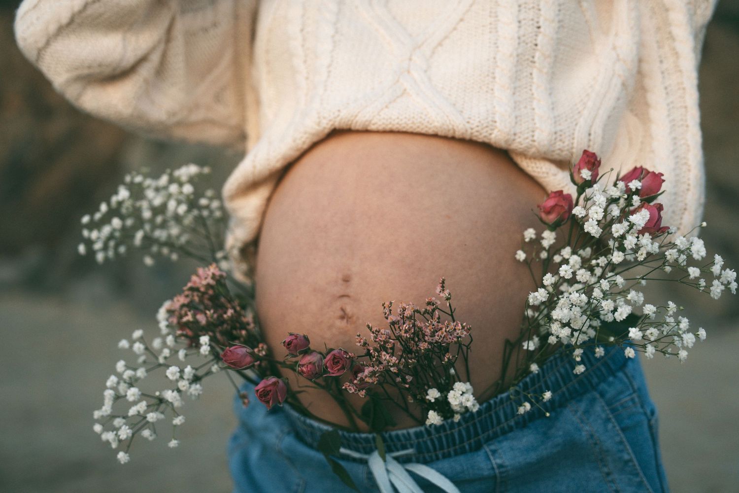 Pregnant woman in white sweater and jeans with wildflowers tucked in waistband showcasing baby bump.