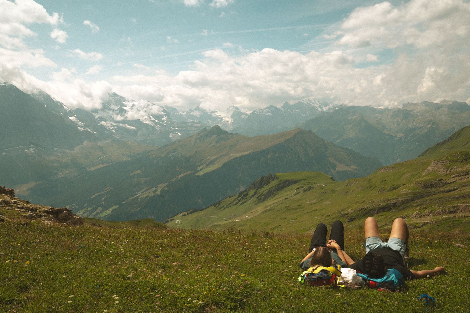Two people relax on a grassy mountain slope overlooking snow-capped peaks and valleys in an alpine landscape.
