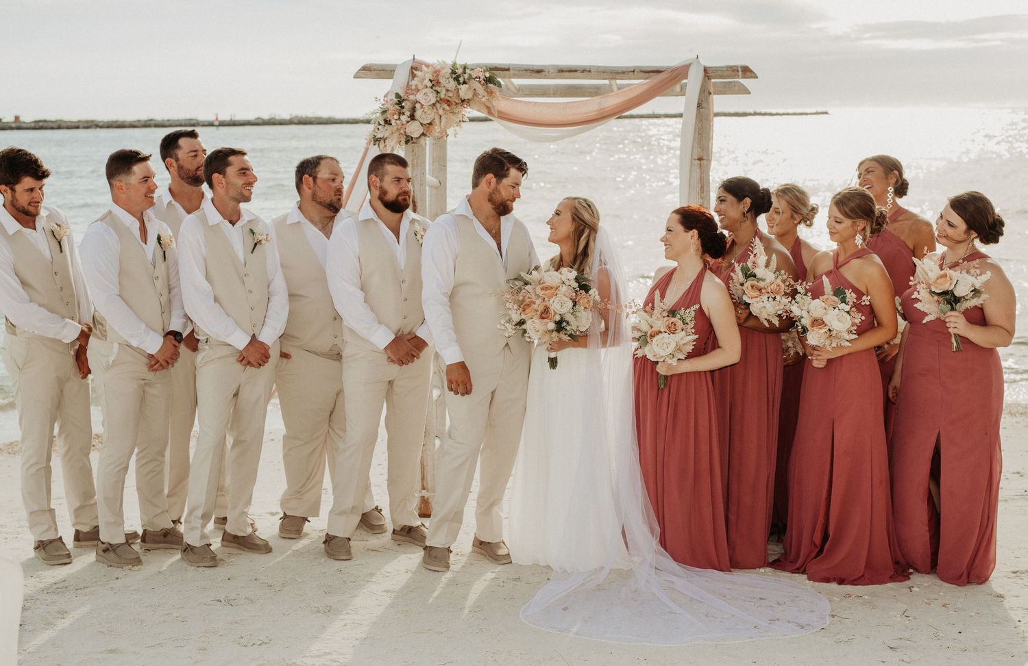 Bridal party in white and terracotta dresses poses together on sandy beach for elegant wedding photos.