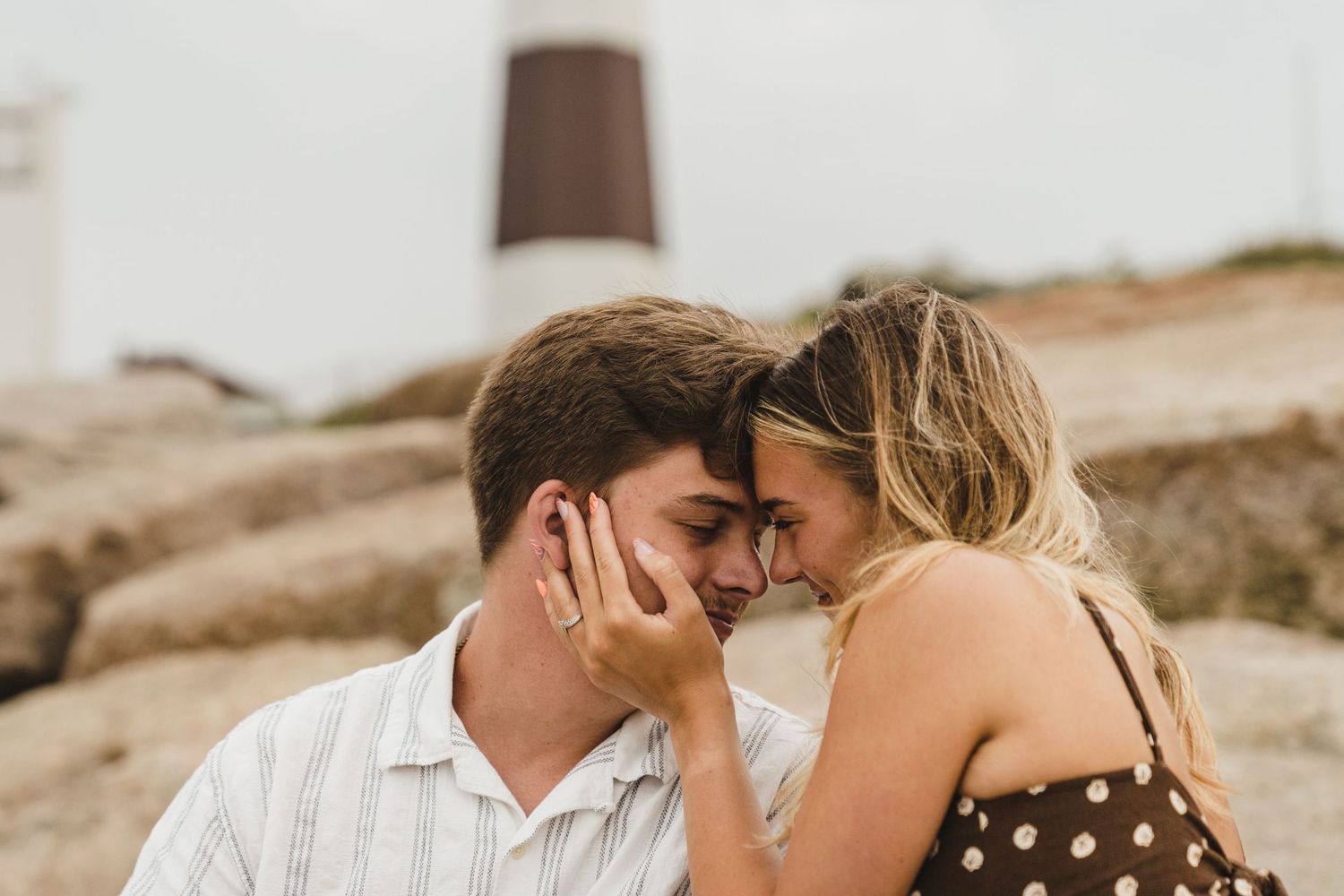 Couple sharing intimate moment on rocky beach landscape during golden hour with woman in polka dot dress.