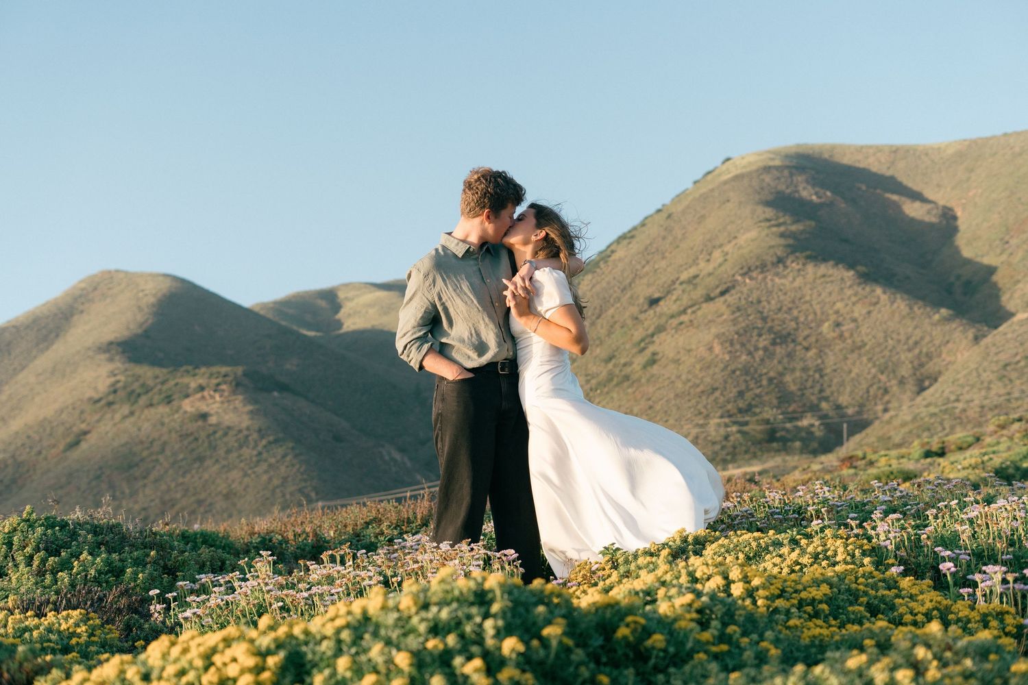 Couple sharing a romantic kiss in a wildflower meadow with rolling green hills and clear blue sky backdrop.