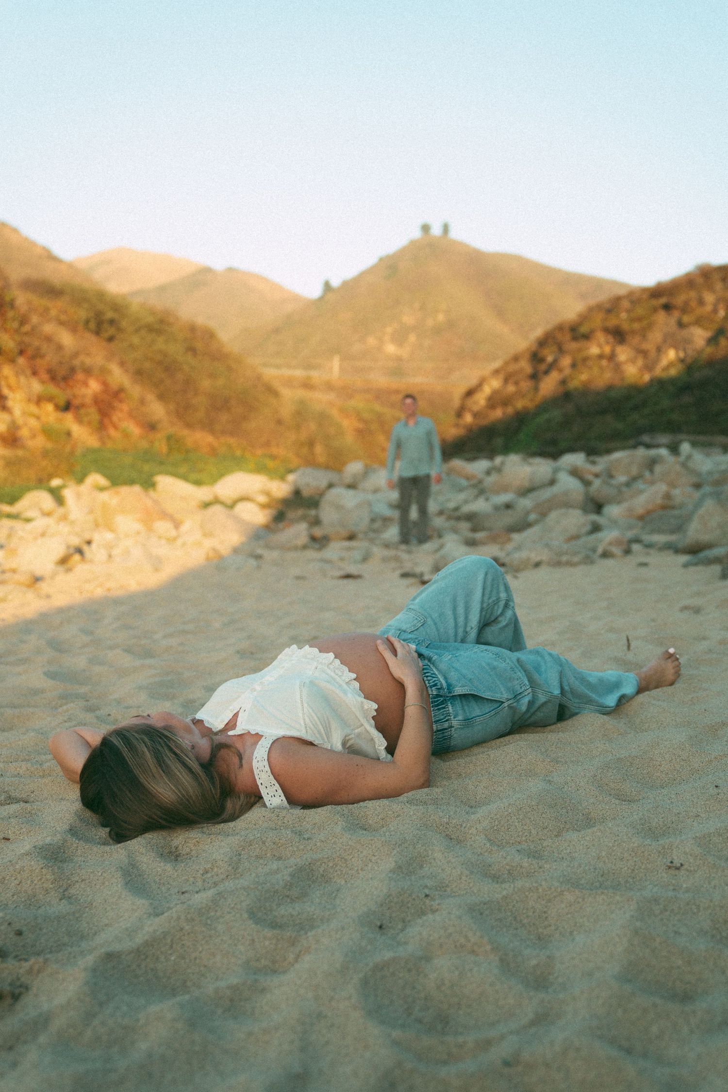 Person lying on sandy beach at sunset with mountains and rolling hills in background during golden hour.