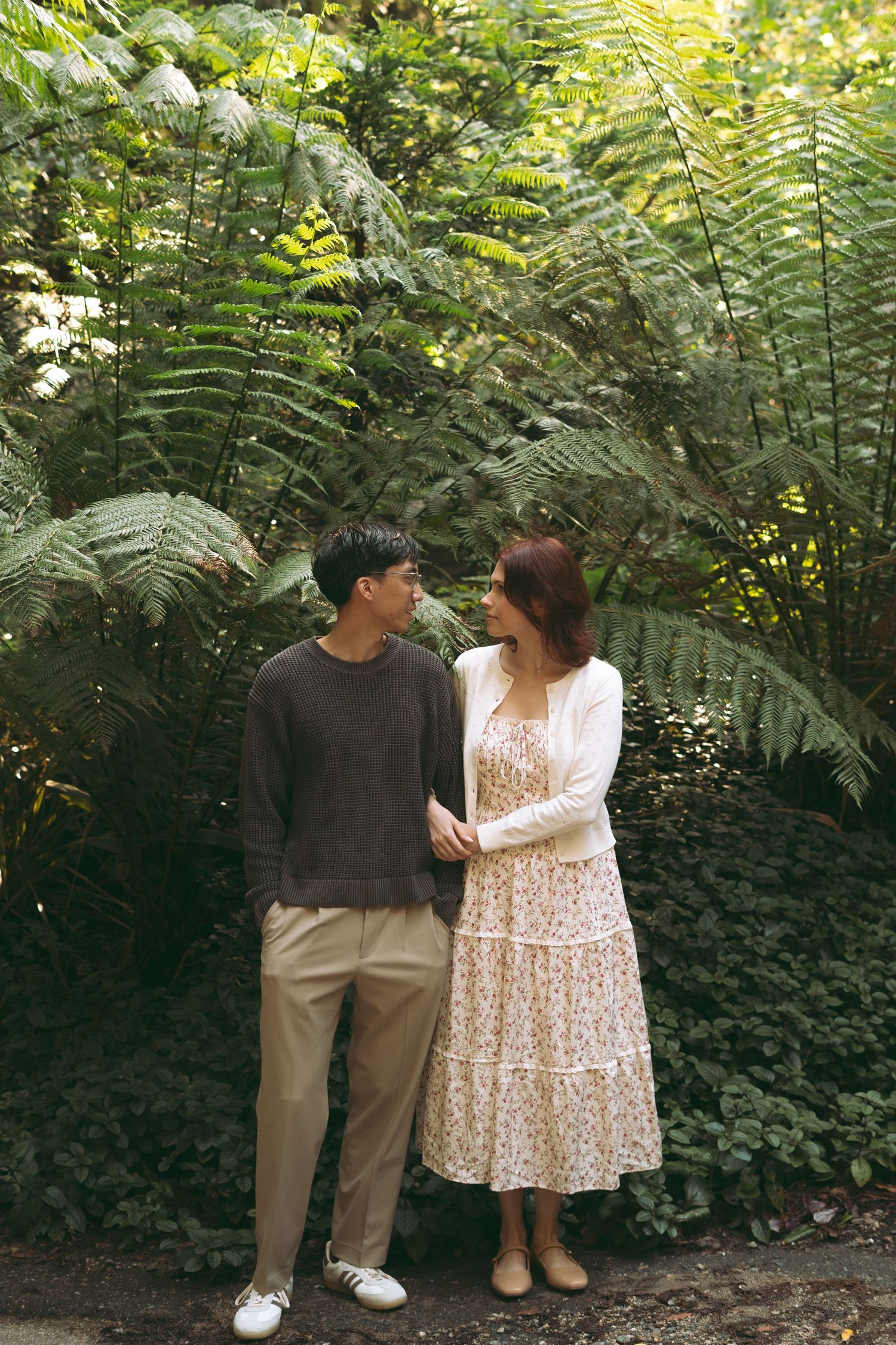 Couple standing together in a lush green garden surrounded by large ferns, smiling at each other.