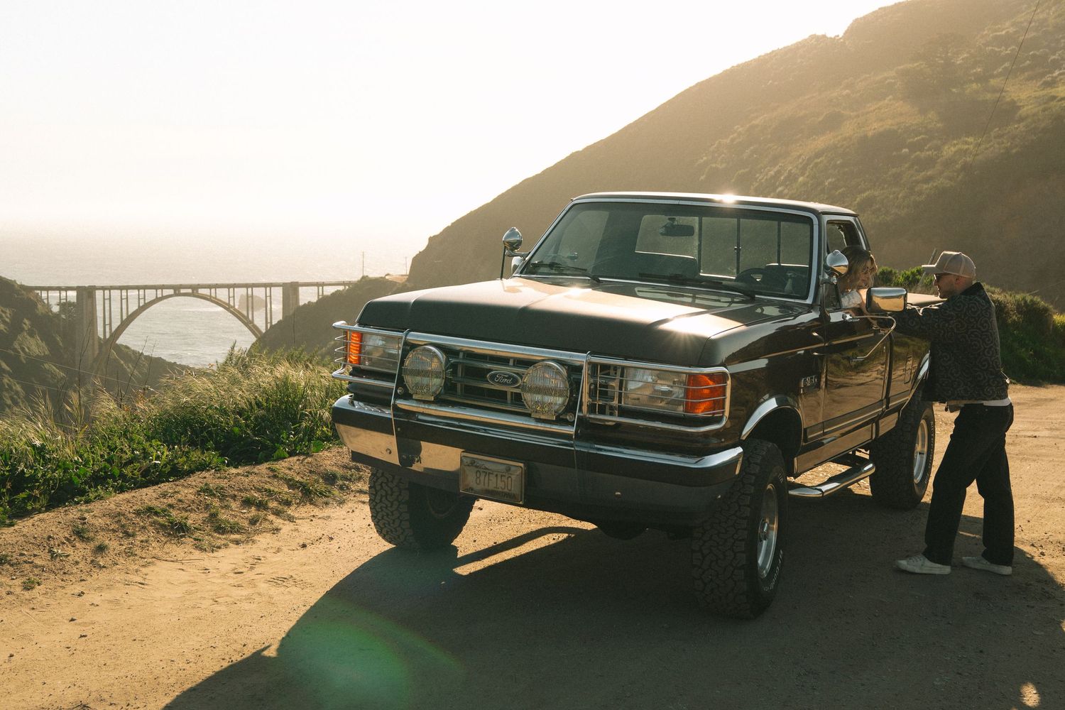 Vintage black Ford truck parked on coastal dirt road near Bixby Bridge, Big Sur, California at golden hour.