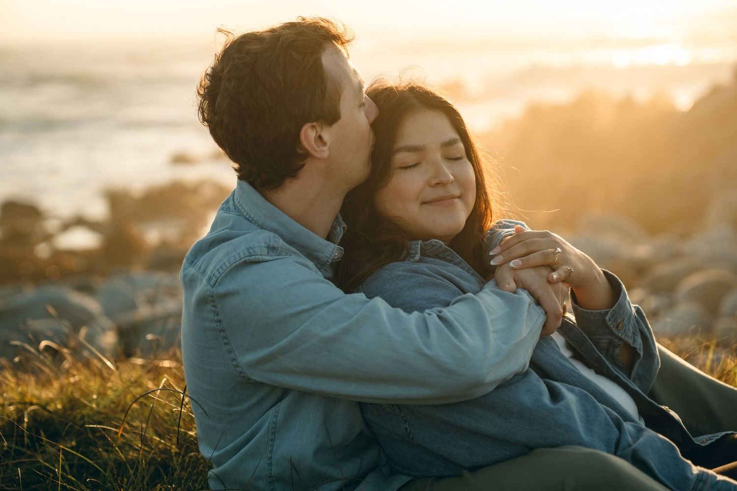 Couple sharing romantic moment while sitting in coastal grass at sunset.