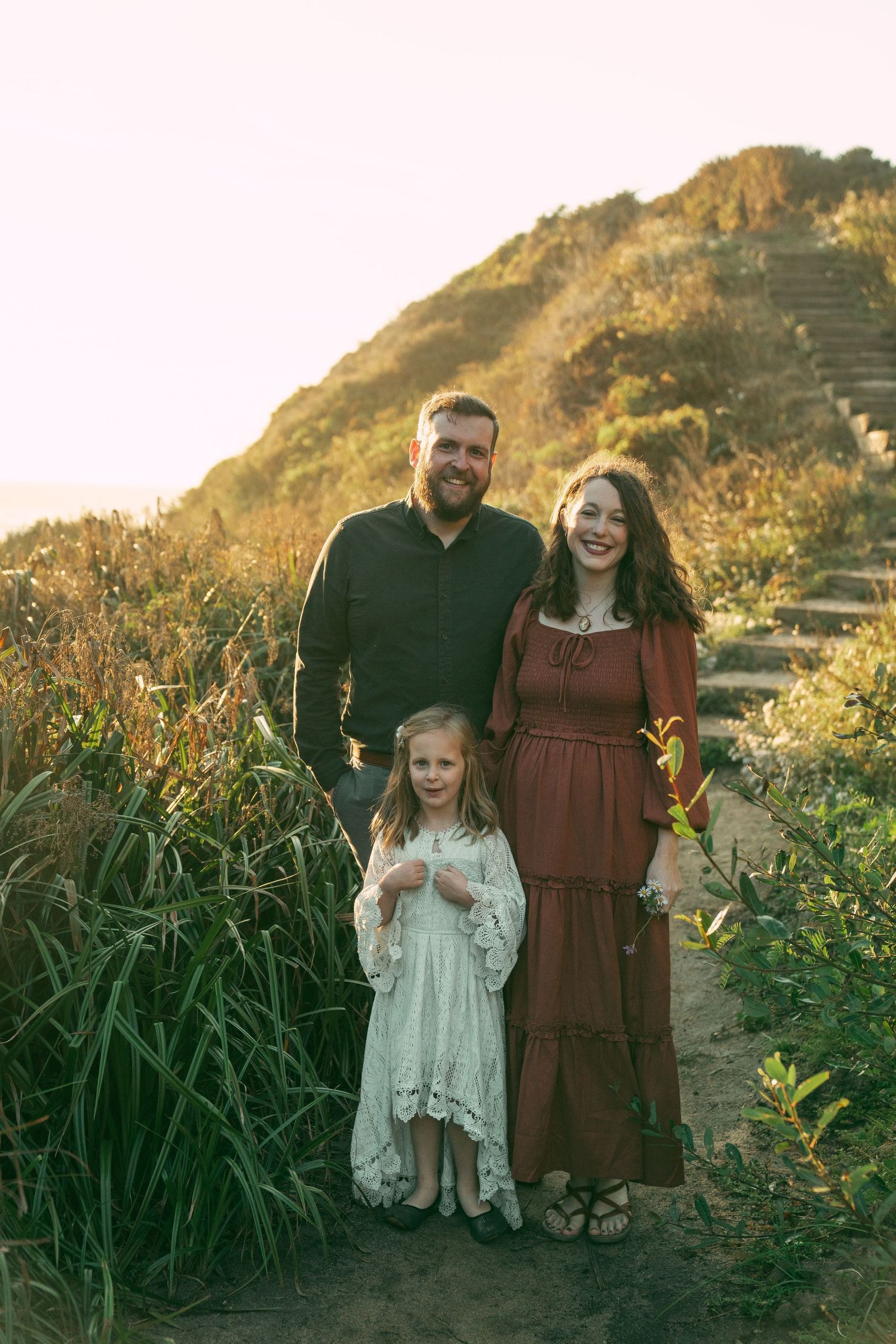 A family poses together at sunset among tall grasses and wildflowers on a coastal hillside.