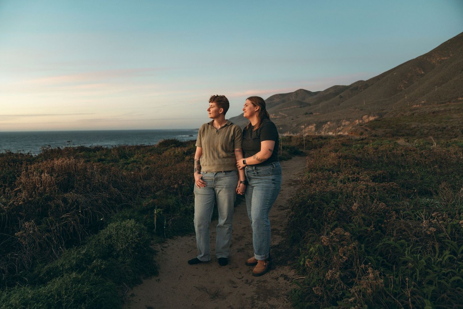 Two people sitting on coastal rocks at sunset overlooking the ocean and rugged coastline.