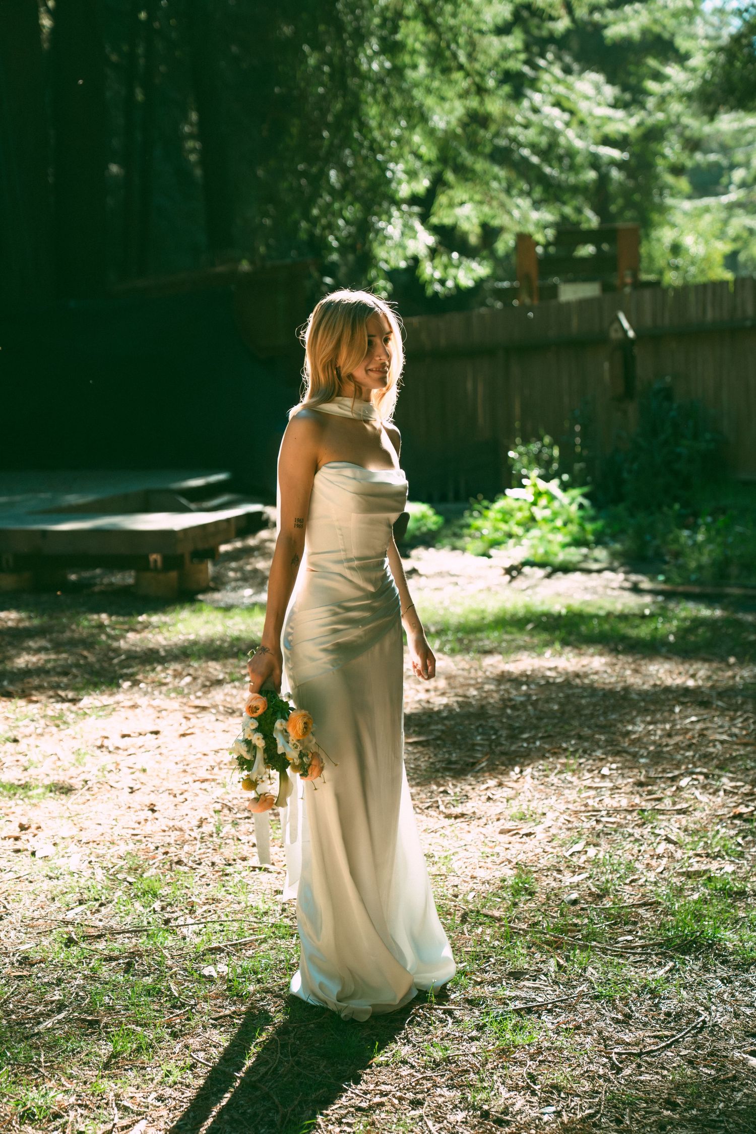 Woman in flowing white dress walking through a sunlit wooded area holding flowers.