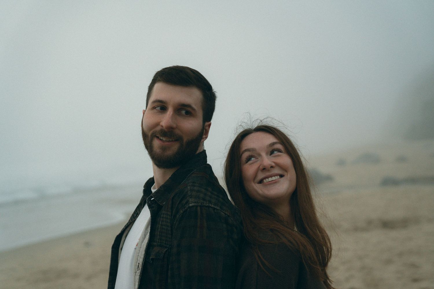 A couple poses together on a misty beach with dramatic coastal scenery.