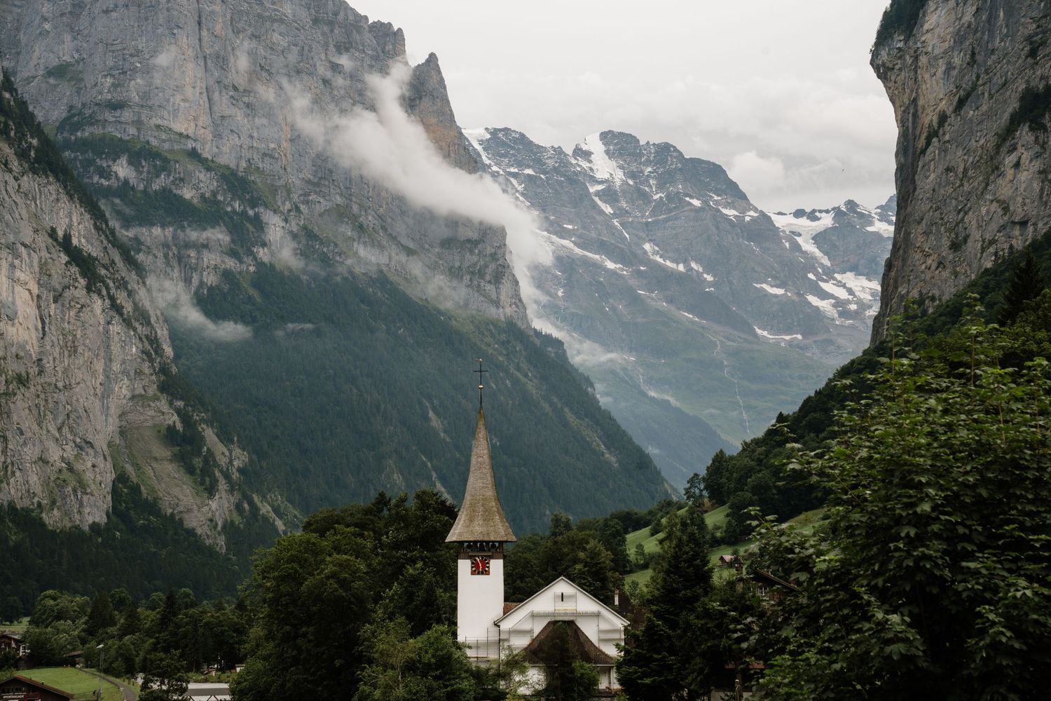 Small white church with spire nestled beneath dramatic snow-capped mountains in Swiss Alps valley on misty day.