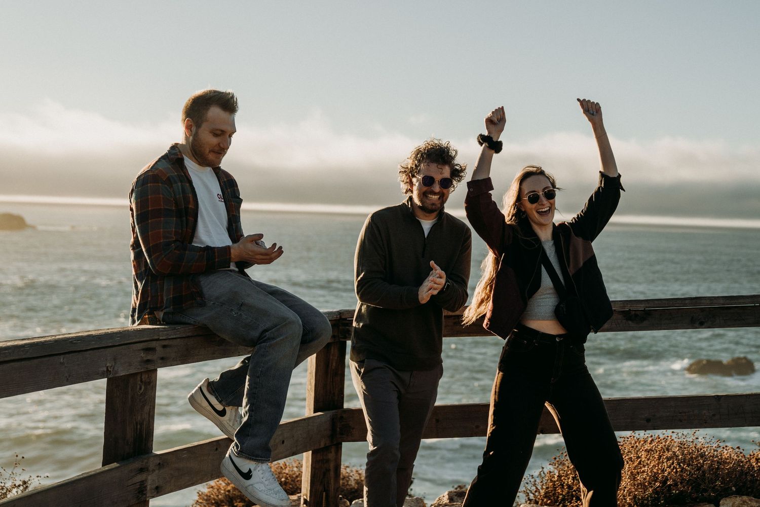 Friends celebrate and pose together on a wooden railing by the ocean at sunset.