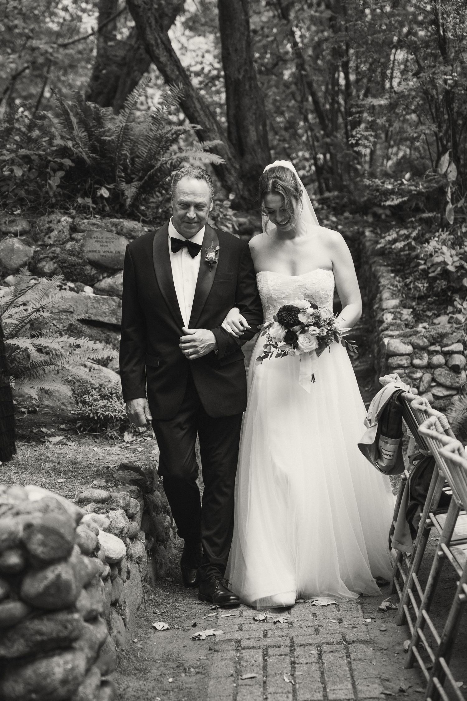 A wedding ceremony procession on a rustic brick path surrounded by lush greenery and stone walls.