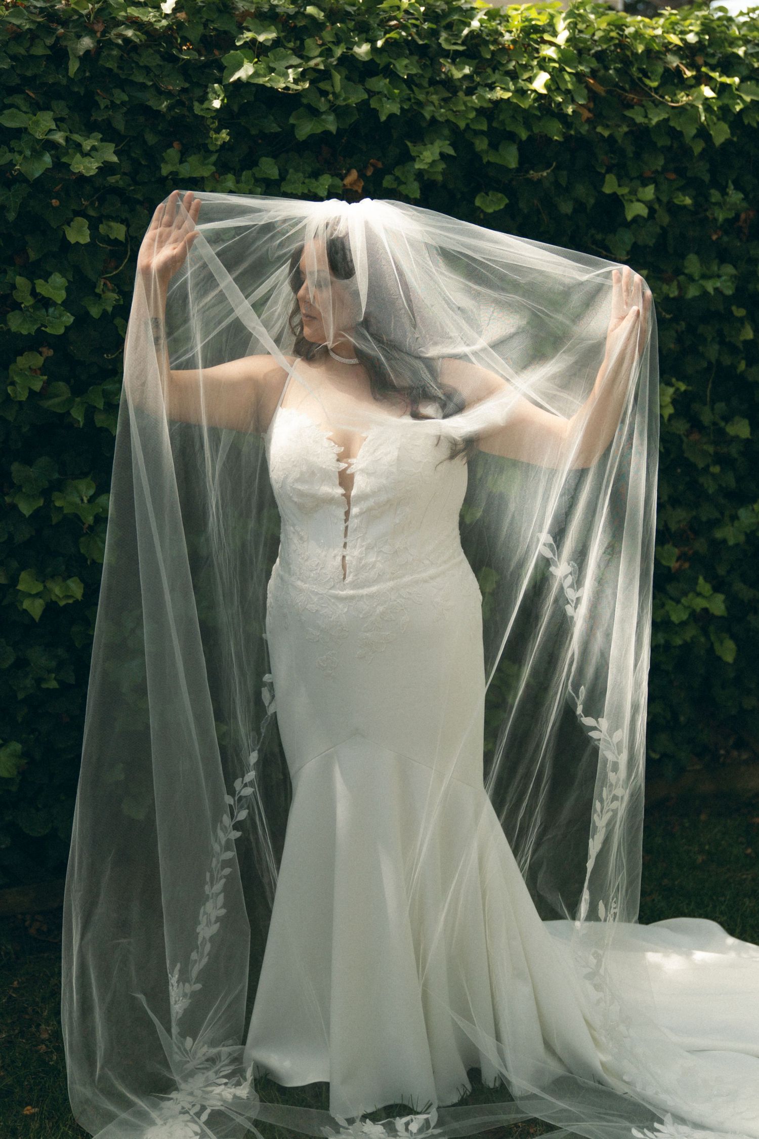 A bride in a flowing white wedding dress and long veil poses gracefully against a backdrop of lush green foliage.