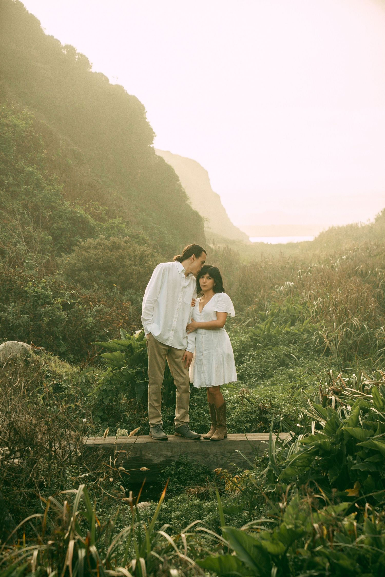 A couple shares an intimate moment on a wooden platform surrounded by lush mountain greenery at sunset.
