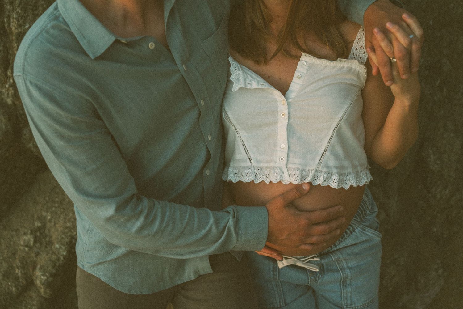 Romantic couple in teal and white clothing embraces intimately against dark textured background during photoshoot.