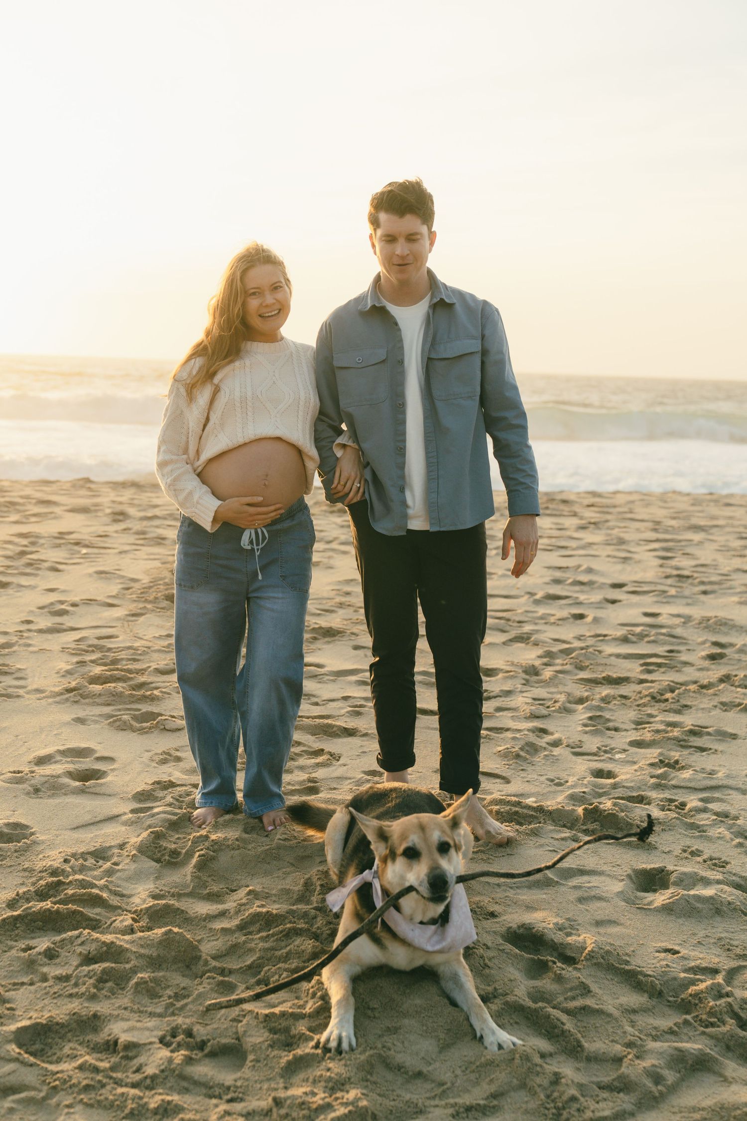 Pregnant couple holding hands on sandy beach at sunset with their leashed beagle dog sitting in front of them.