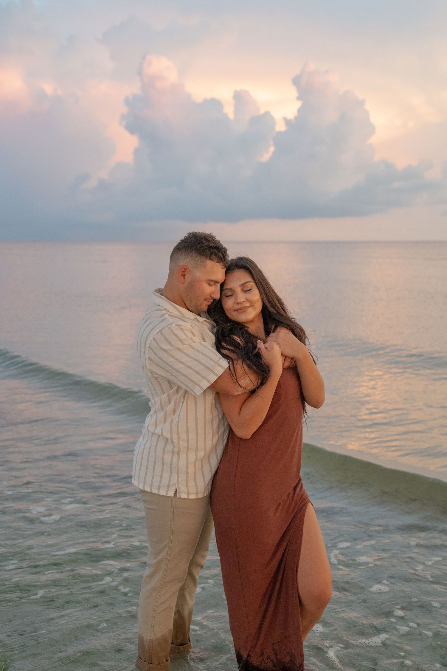 A couple embraces on the beach during golden hour with pastel clouds and ocean in the background.