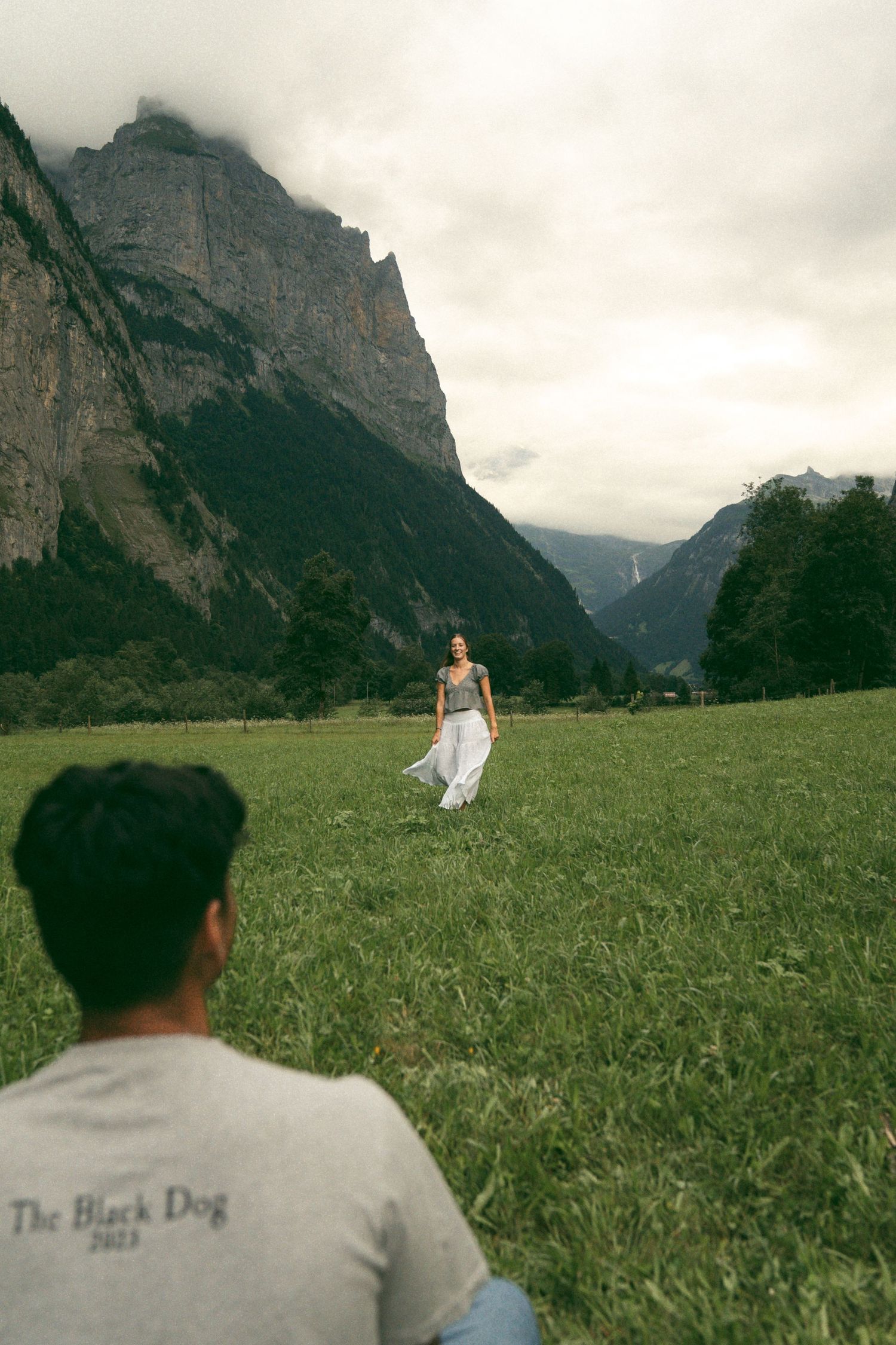 A person in white stands in a grassy meadow with dramatic mountain peaks rising in the background under cloudy skies.