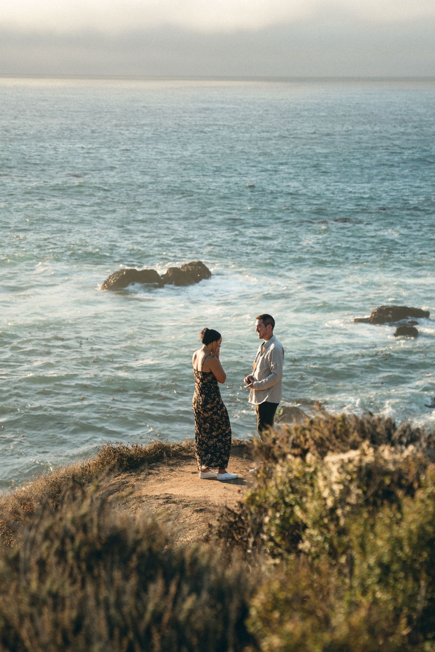 A couple stands on a coastal cliff overlooking the ocean during golden hour with waves crashing against rocks below.