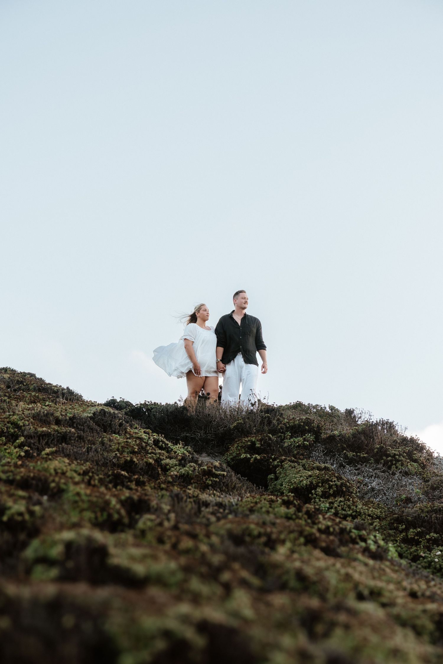 A couple and their dog stand together on a rocky hilltop overlooking the landscape against a light blue sky.