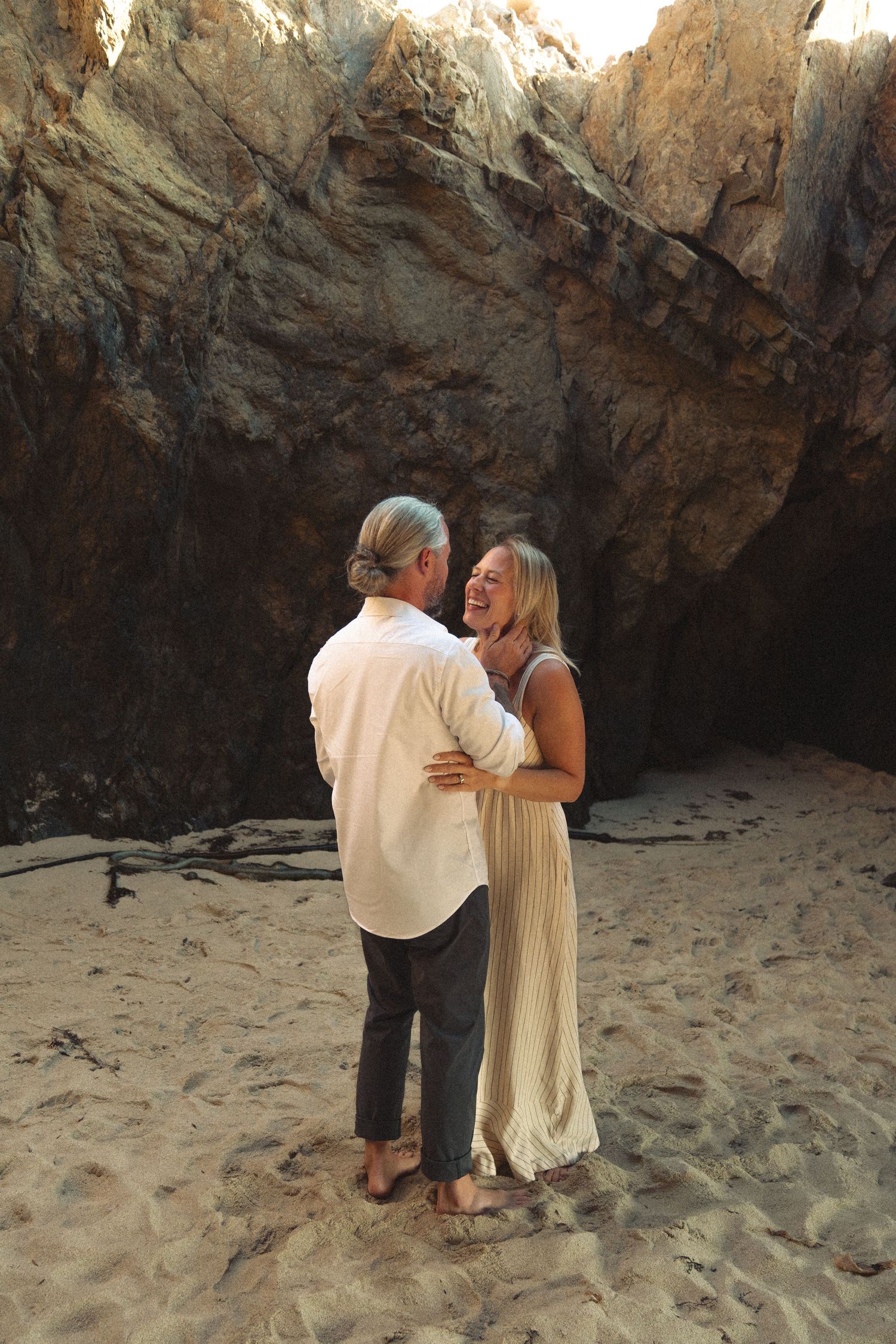 A couple shares an intimate moment on a secluded beach near rocky cliffs during sunset.