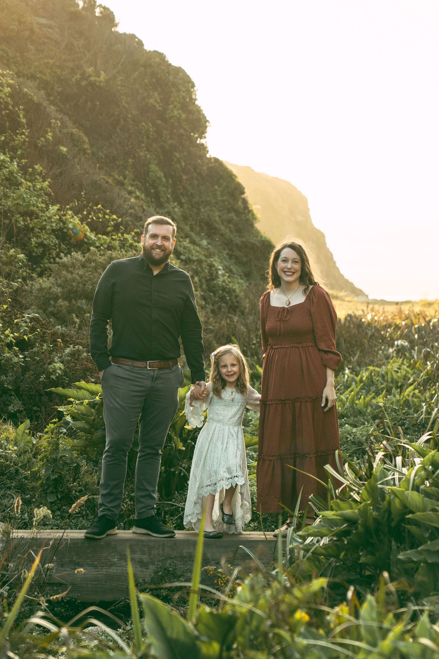A family poses together on a coastal path surrounded by green foliage and mountainous cliffs at sunset.