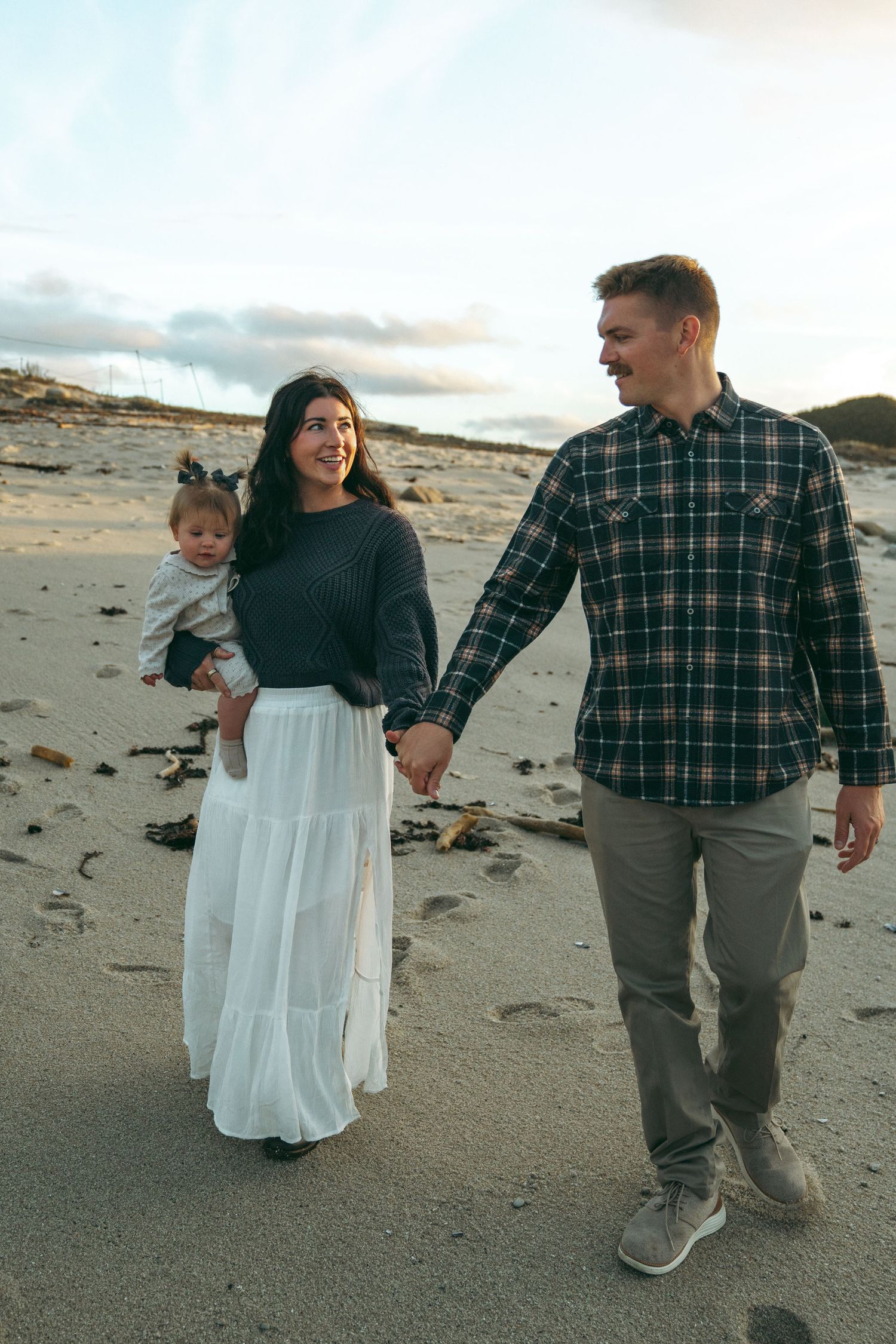 A family takes a casual stroll along a sandy beach at sunset, wearing coordinated plaid and white outfits.