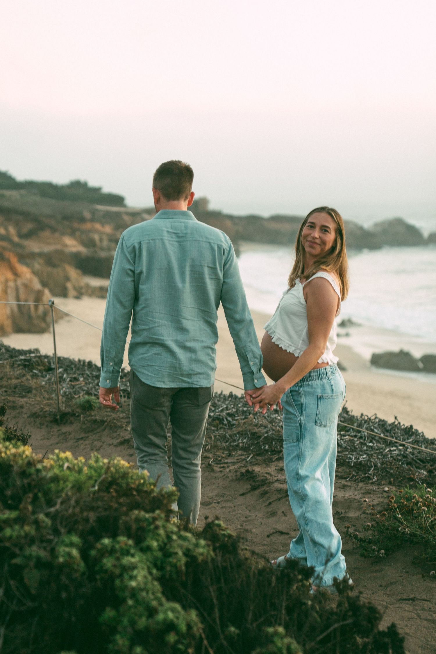 A couple walks along a scenic coastal path at sunset with ocean views and natural vegetation in the foreground.