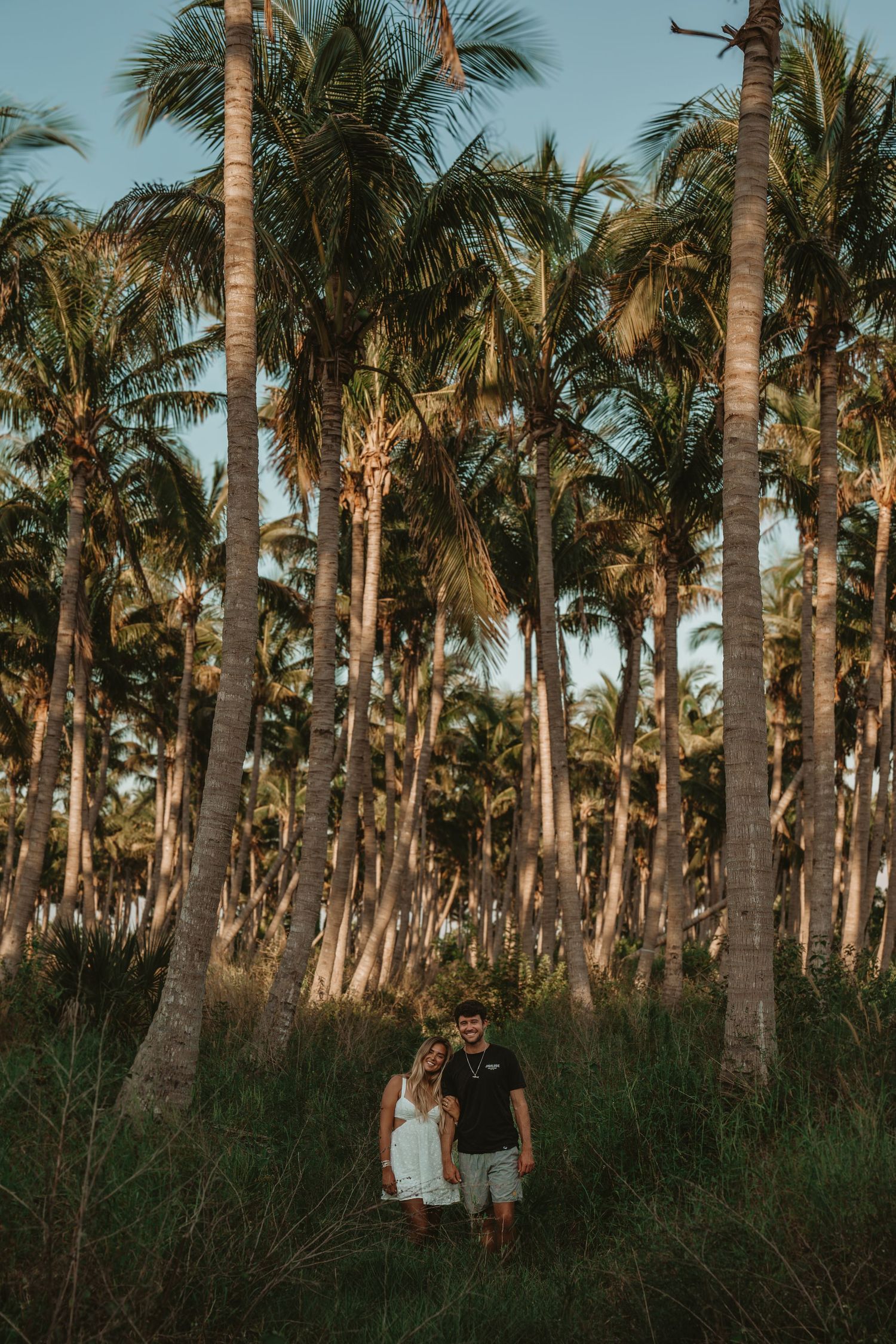 A couple walking through a scenic palm tree grove at sunset casting long shadows through the tropical landscape.