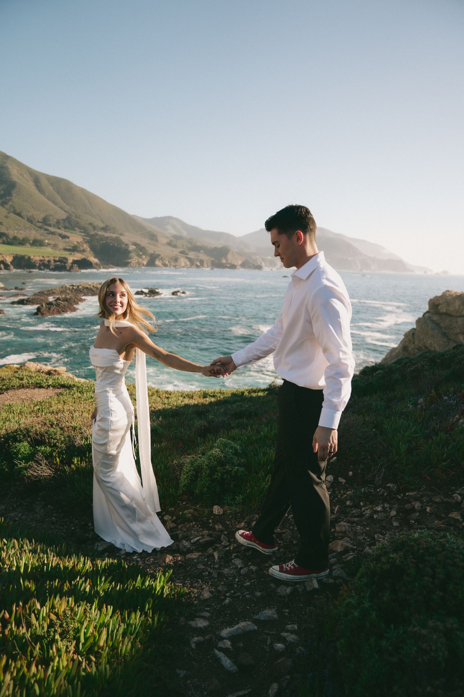 Couple dancing and twirling together on a scenic coastal cliff with mountains and ocean in the background.