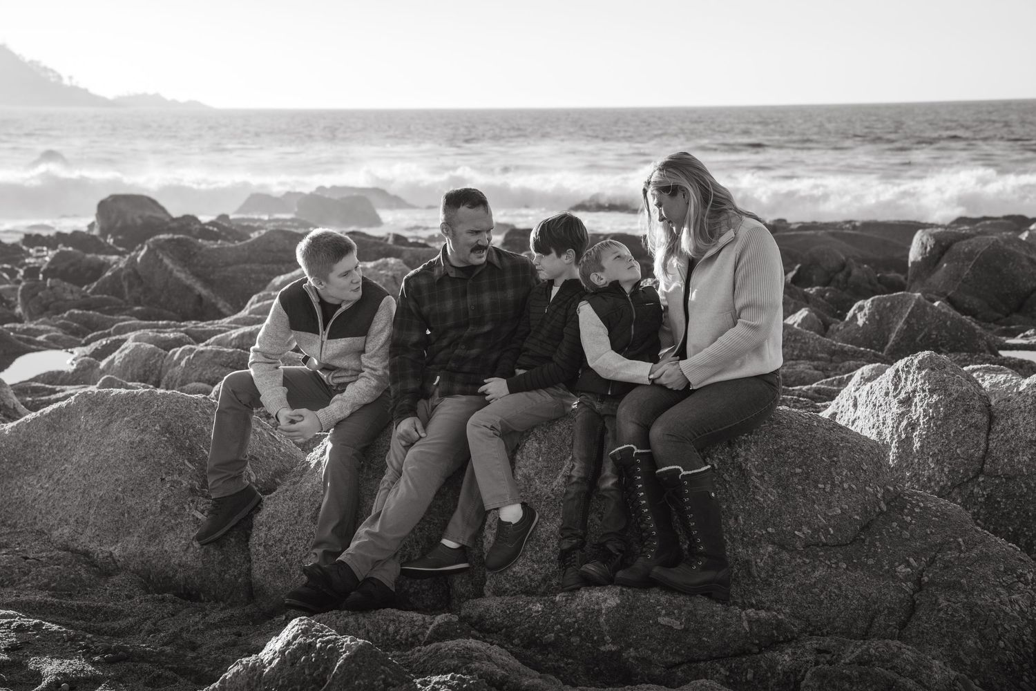 A group sits together on coastal rocks in black and white as ocean waves crash behind them.