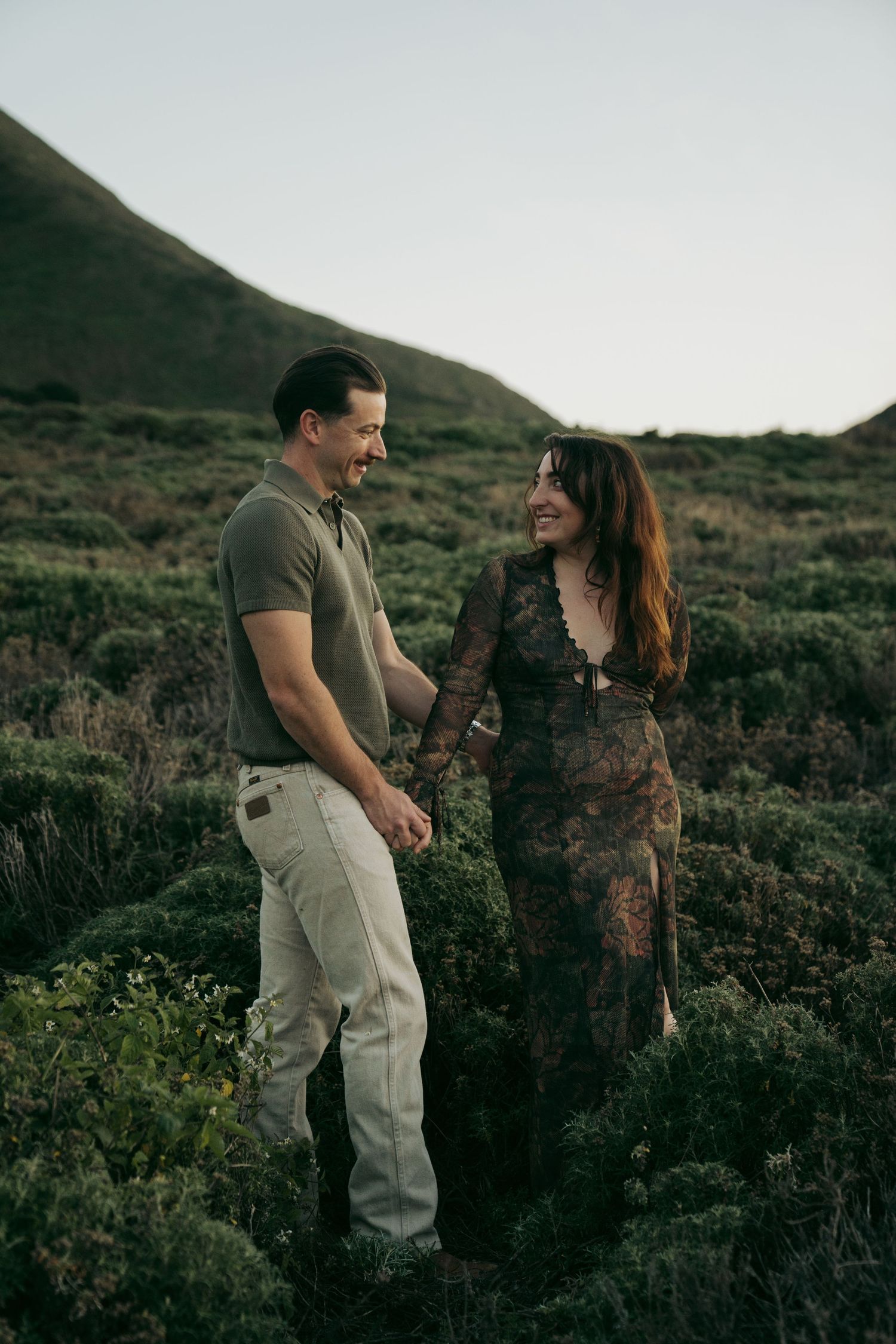 A couple shares a romantic moment while hiking through a scenic mountainous landscape at dusk.