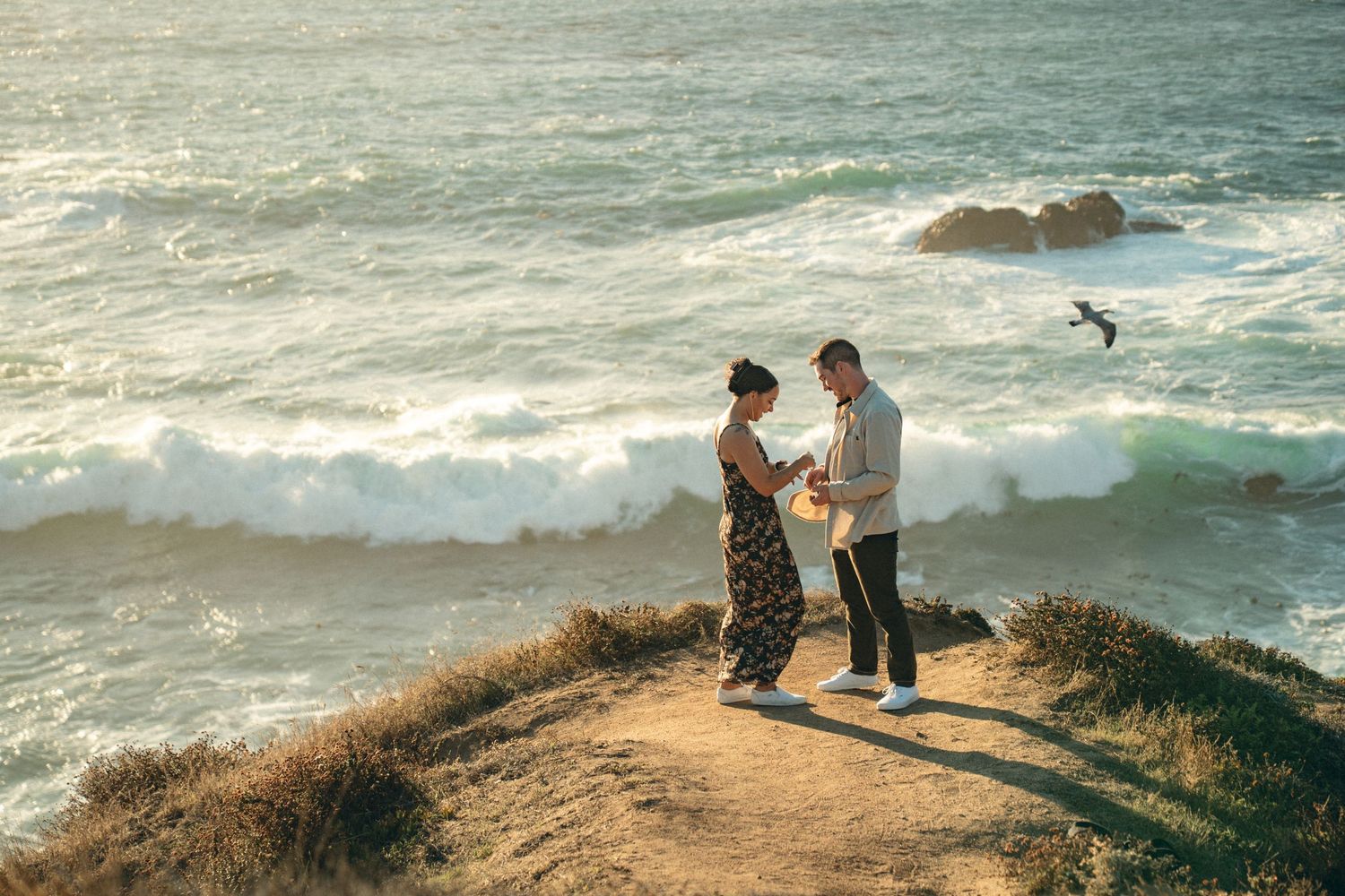 Silhouetted figures stand along a rugged coastal cliff overlooking the crashing waves of the Pacific Ocean at sunset.