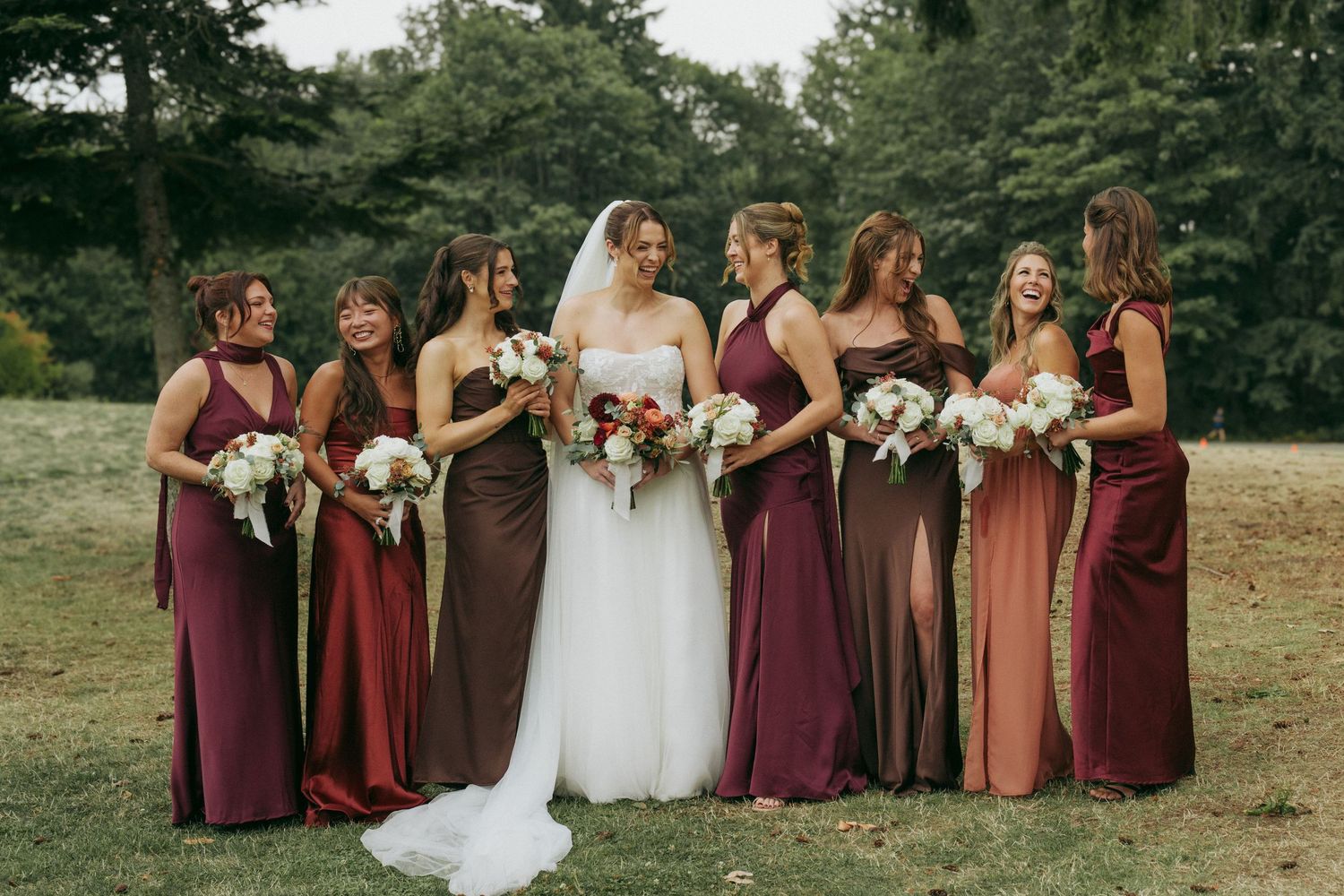 A wedding party wearing burgundy and mauve colored dresses poses together outdoors on a grassy field.