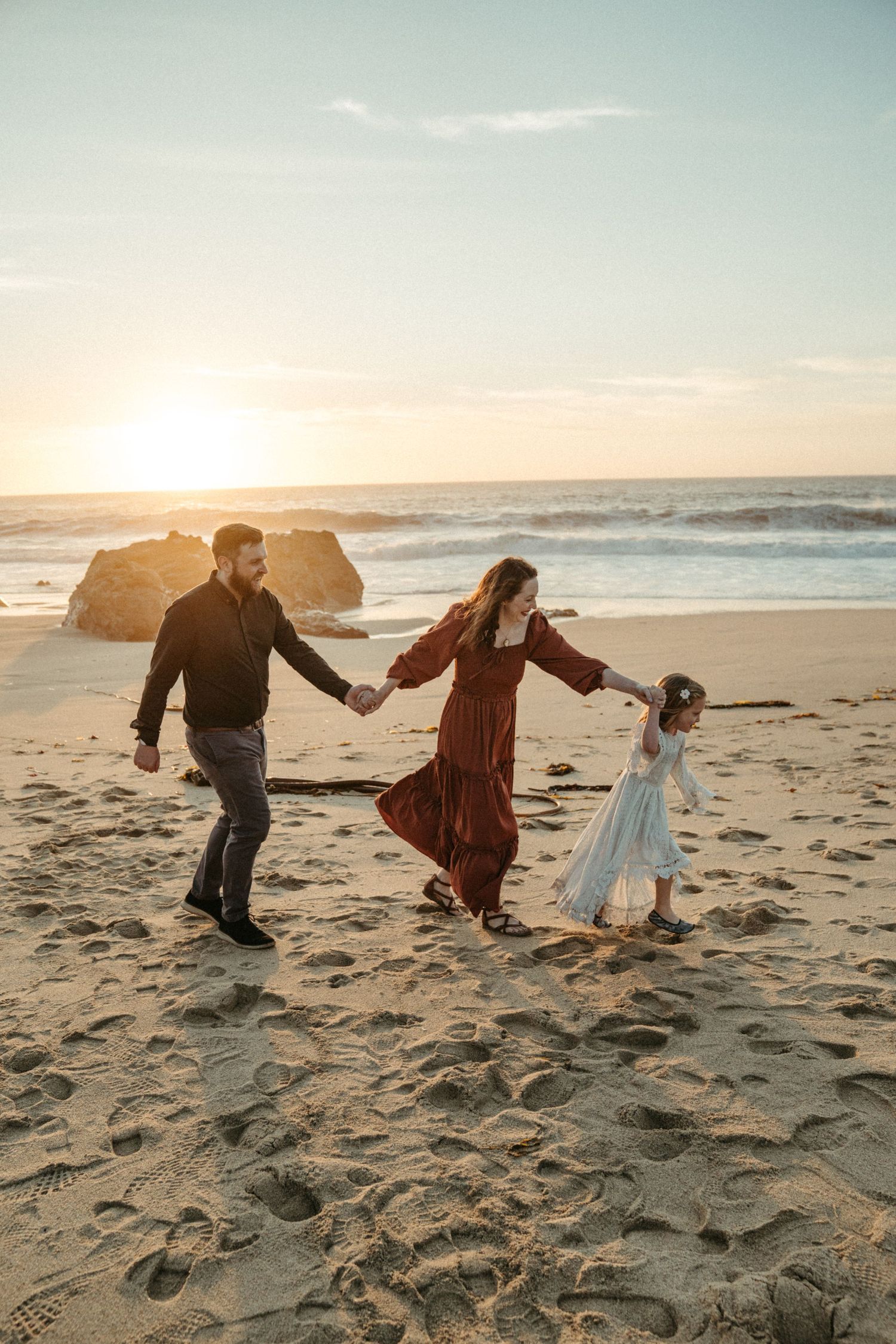 Three people hold hands while walking along a sandy beach at sunset creating silhouettes against the golden sky.