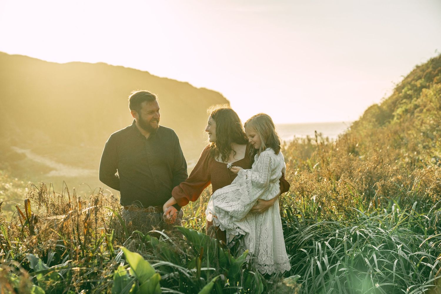 Silhouetted figures sit together on grassy hillside during golden sunset hour with mountains in background.