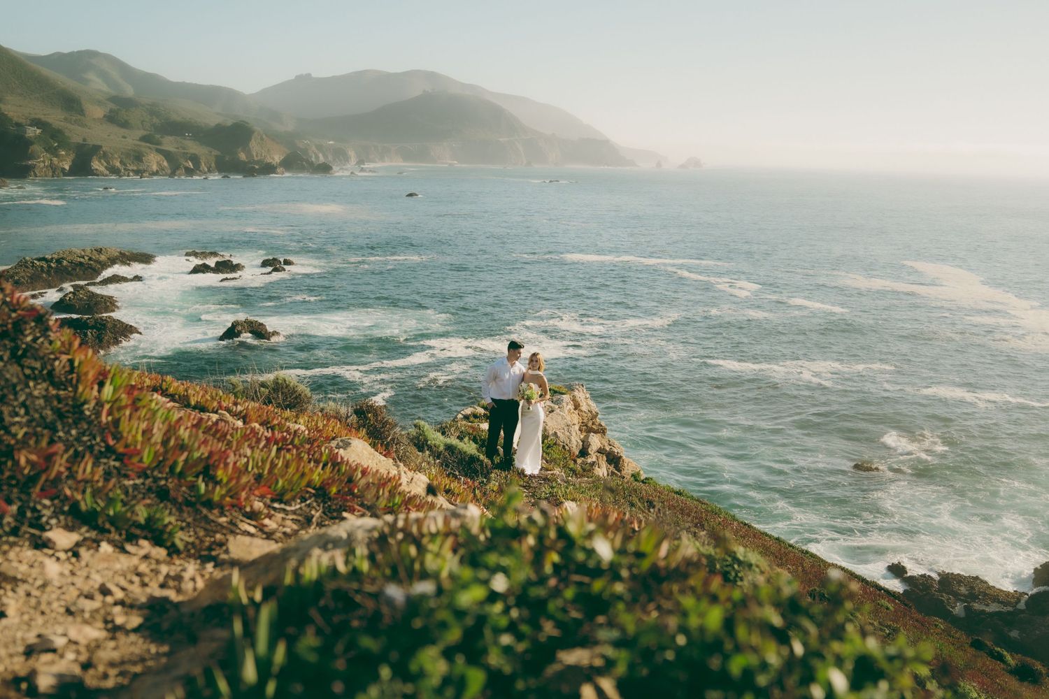 Panoramic view of rugged California coastline with crashing waves against rocky cliffs and misty mountains in background.