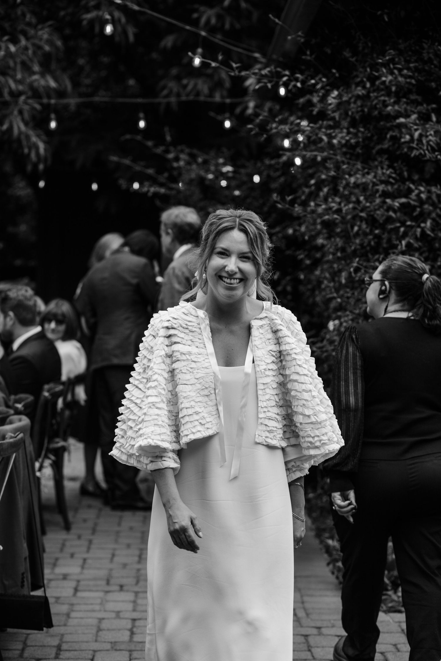 A person in a white wedding dress with a textured shrug stands outdoors at an evening event with string lights.