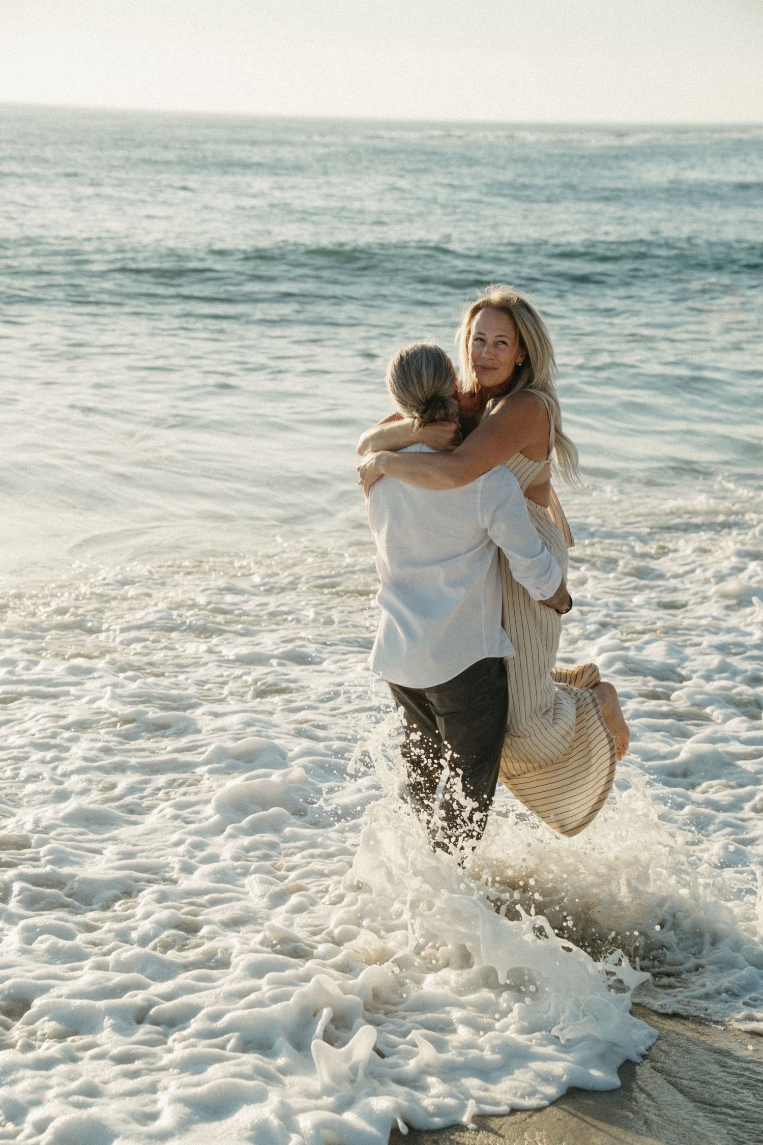A playful couple splashes through ocean waves at the beach during sunset in white casual attire.