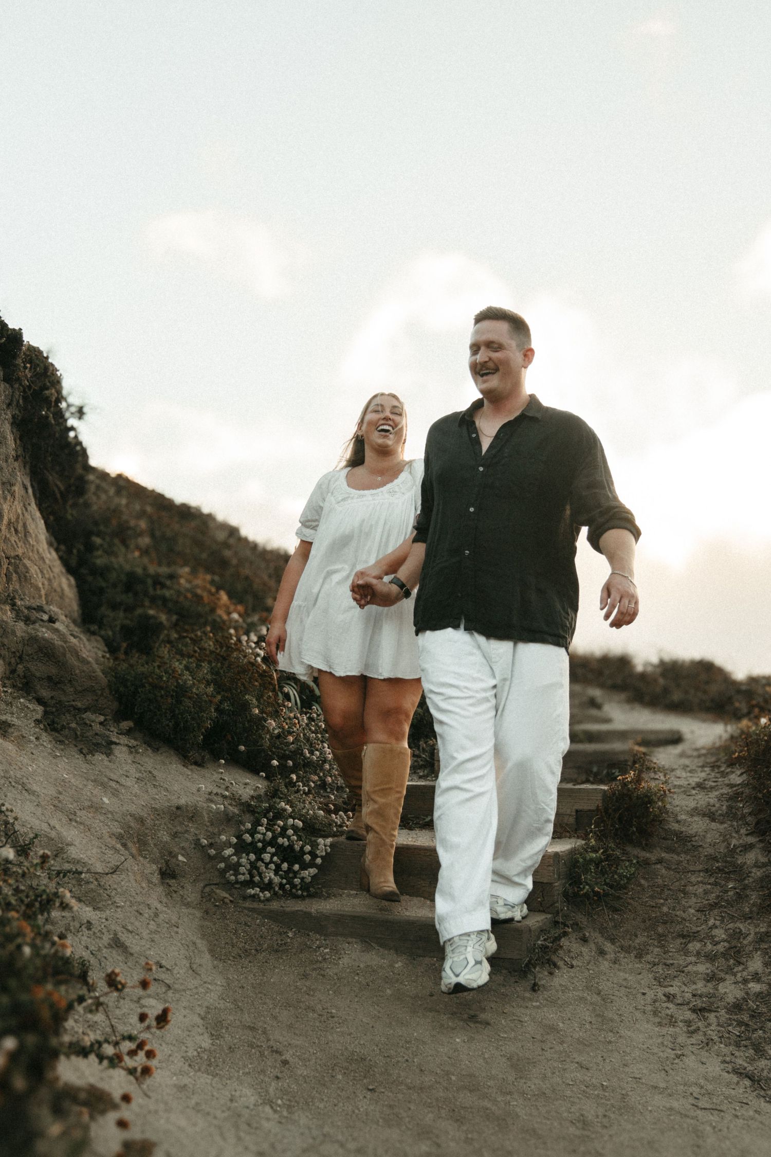 A couple walks together on a scenic coastal path during sunset, one wearing white and the other in dark clothing.