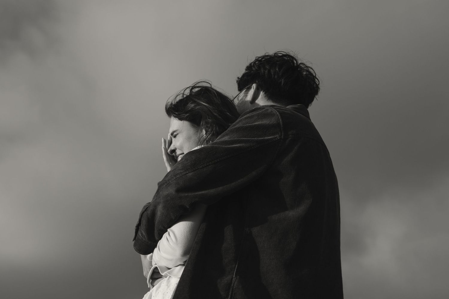 Black and white photo of a couple embracing tenderly under a dramatic cloudy sky.