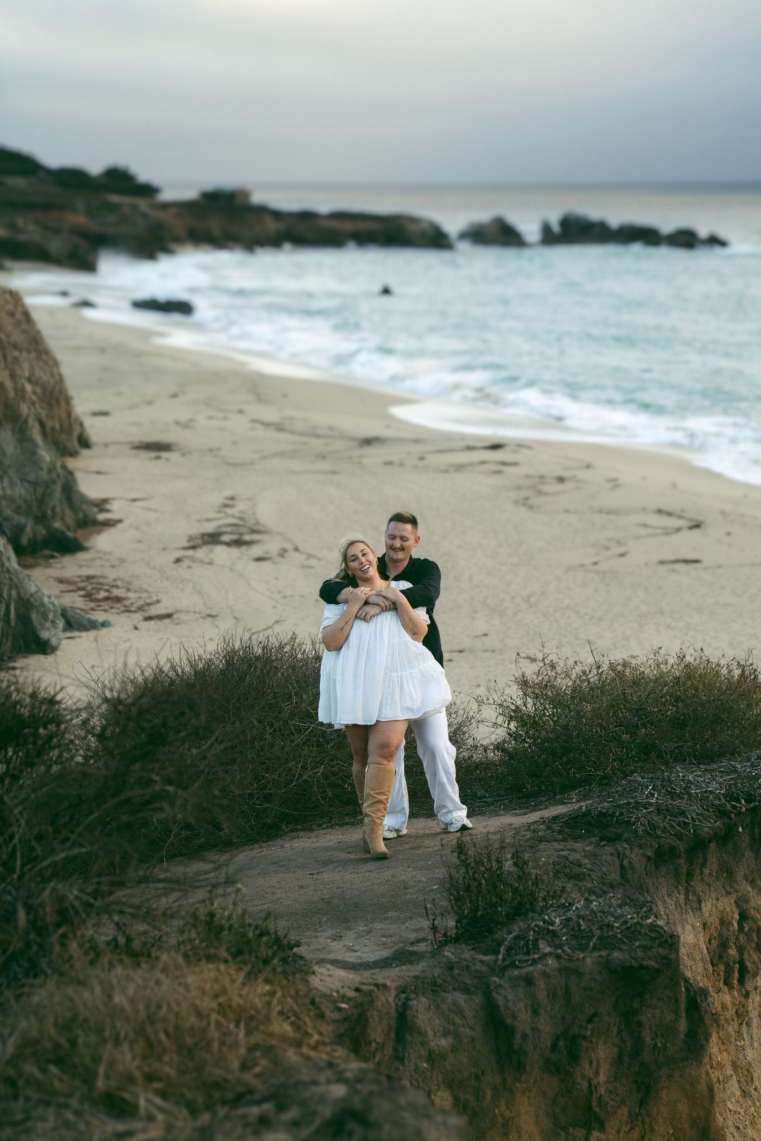 Two people dance together on a dramatic coastal cliff overlooking a beach at dusk.