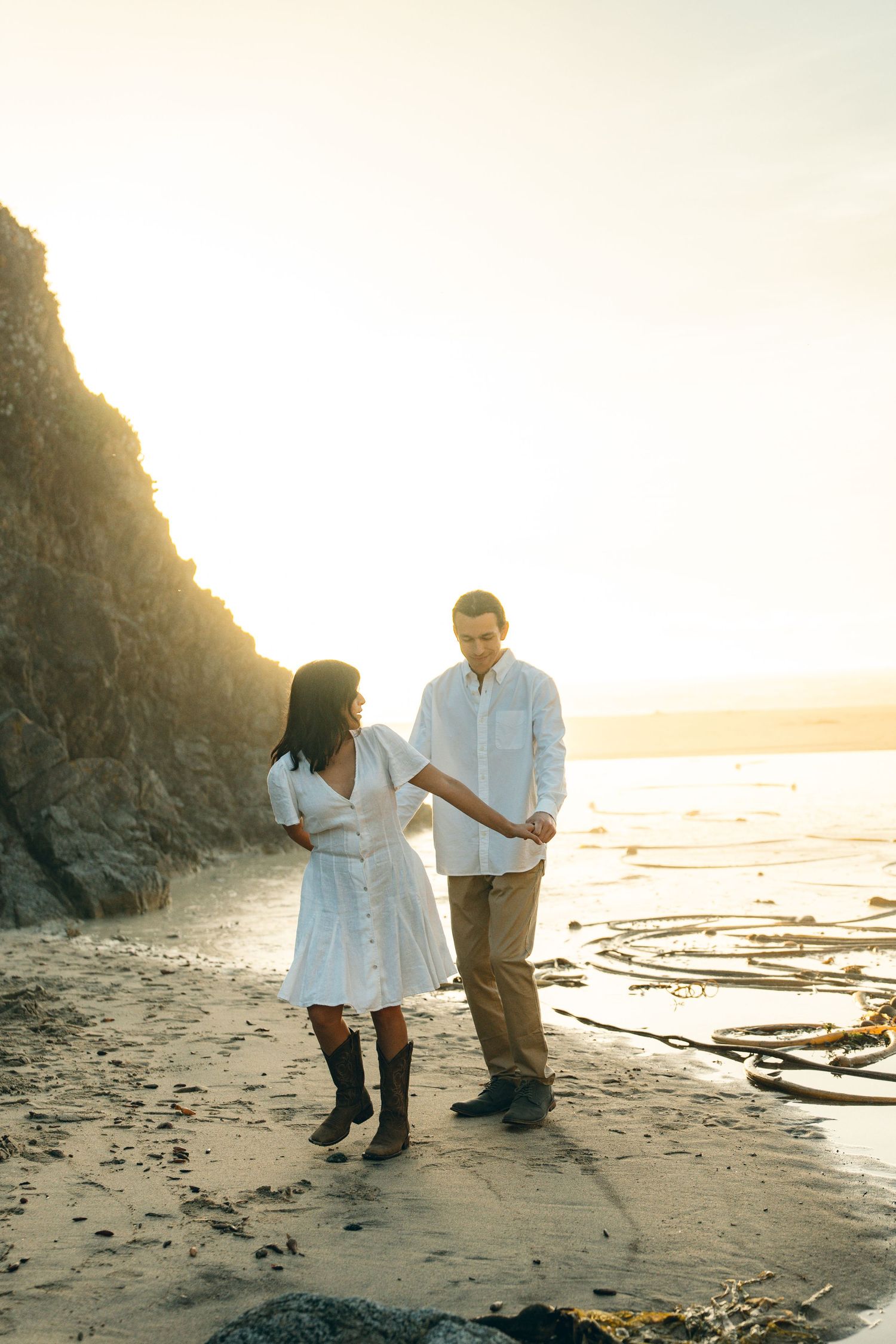 Two people walk along a scenic beach at sunset with cliffs and driftwood in the background.