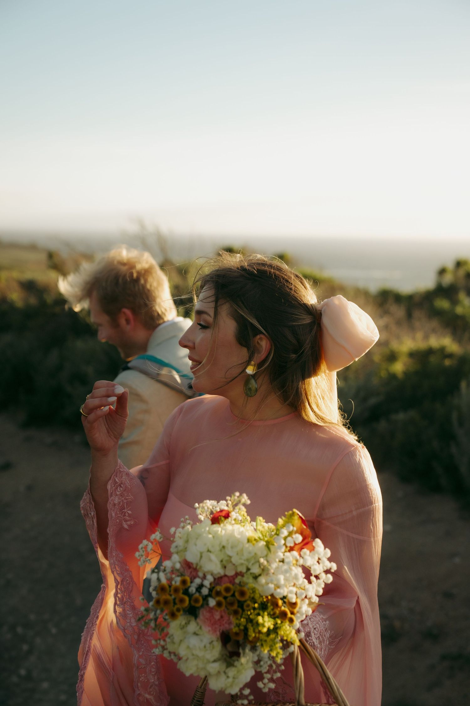 Bride in pink sheer gown holds wildflower bouquet at sunset with groom in coastal landscape.
