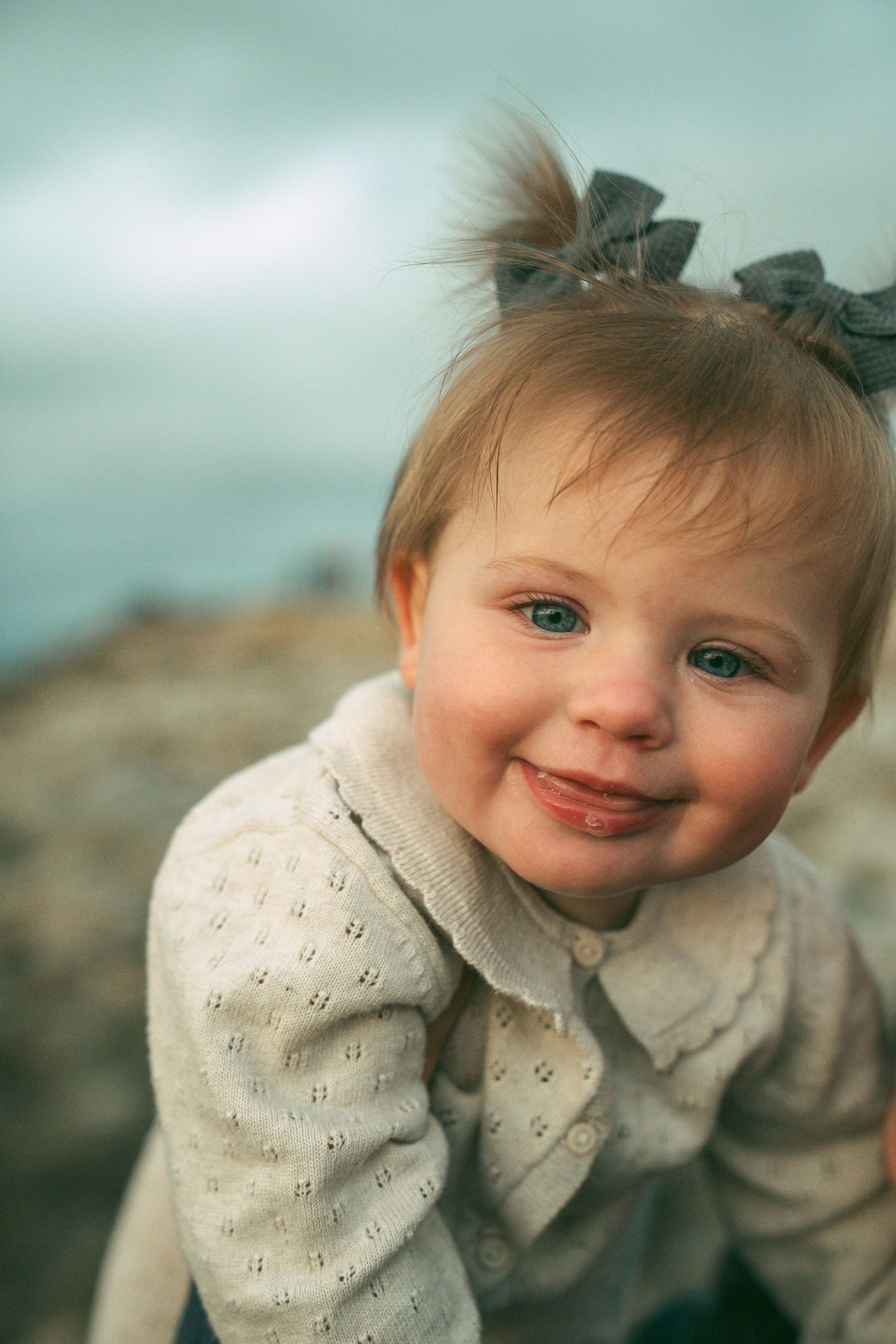 A toddler in a cream-colored eyelet dress and bow smiles sweetly at the camera against a soft blue background.
